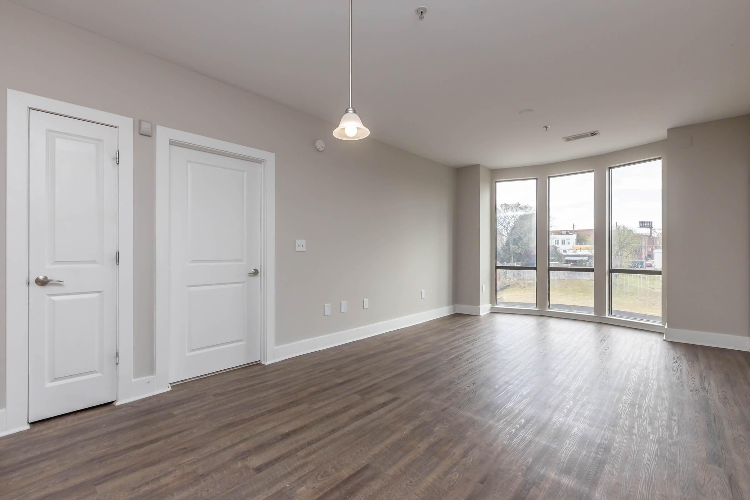 A spacious, empty room with hardwood floors and large windows allowing natural light, featuring two closed doors on the left and a pendant light hanging from the ceiling. The walls are painted in a neutral color, and there is a view of greenery outside the windows.