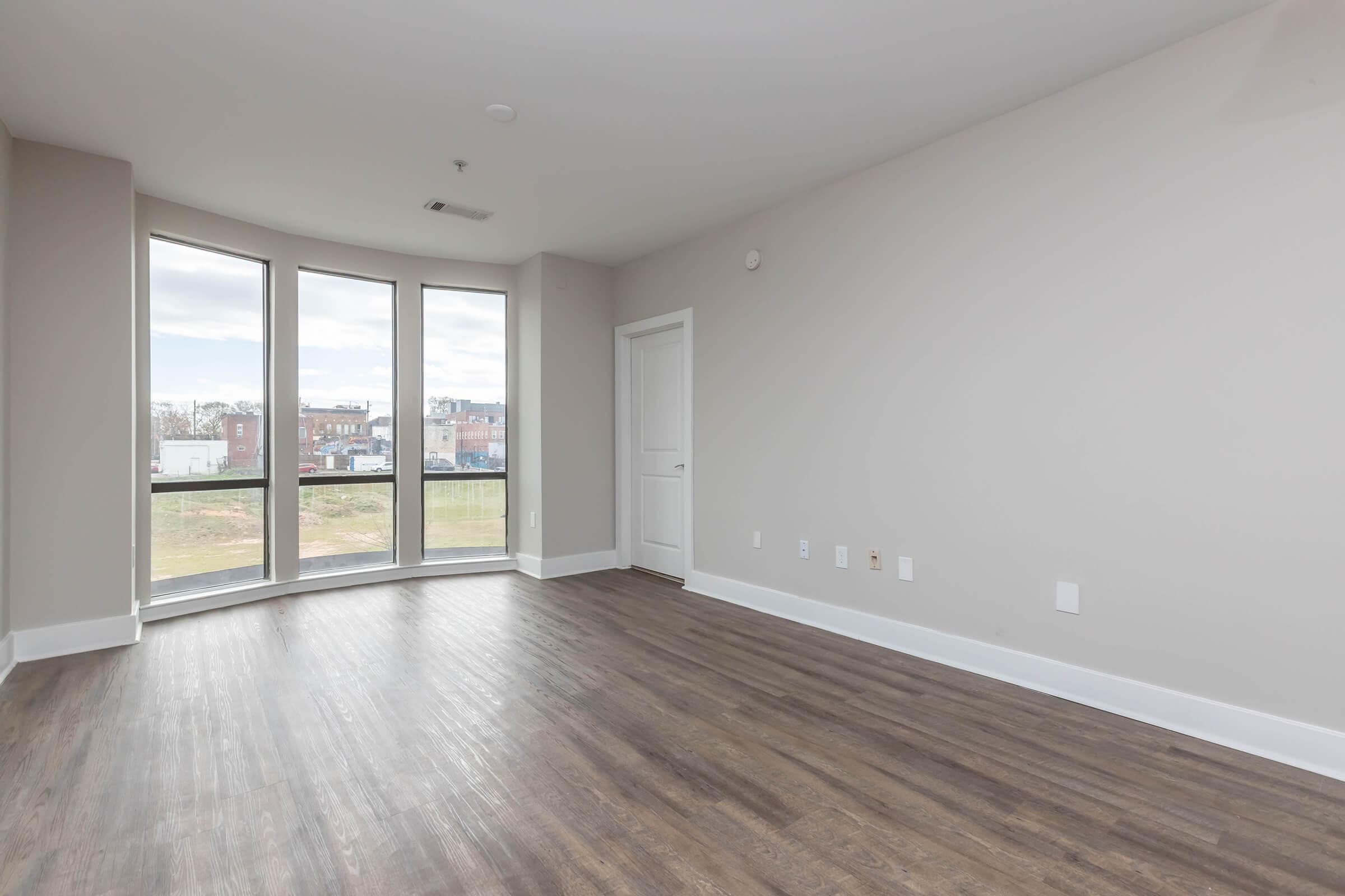 Empty room with hardwood flooring and large windows allowing natural light. The walls are painted in a light color, and there is a single door leading to another room. The view outside features a landscape with nearby buildings.