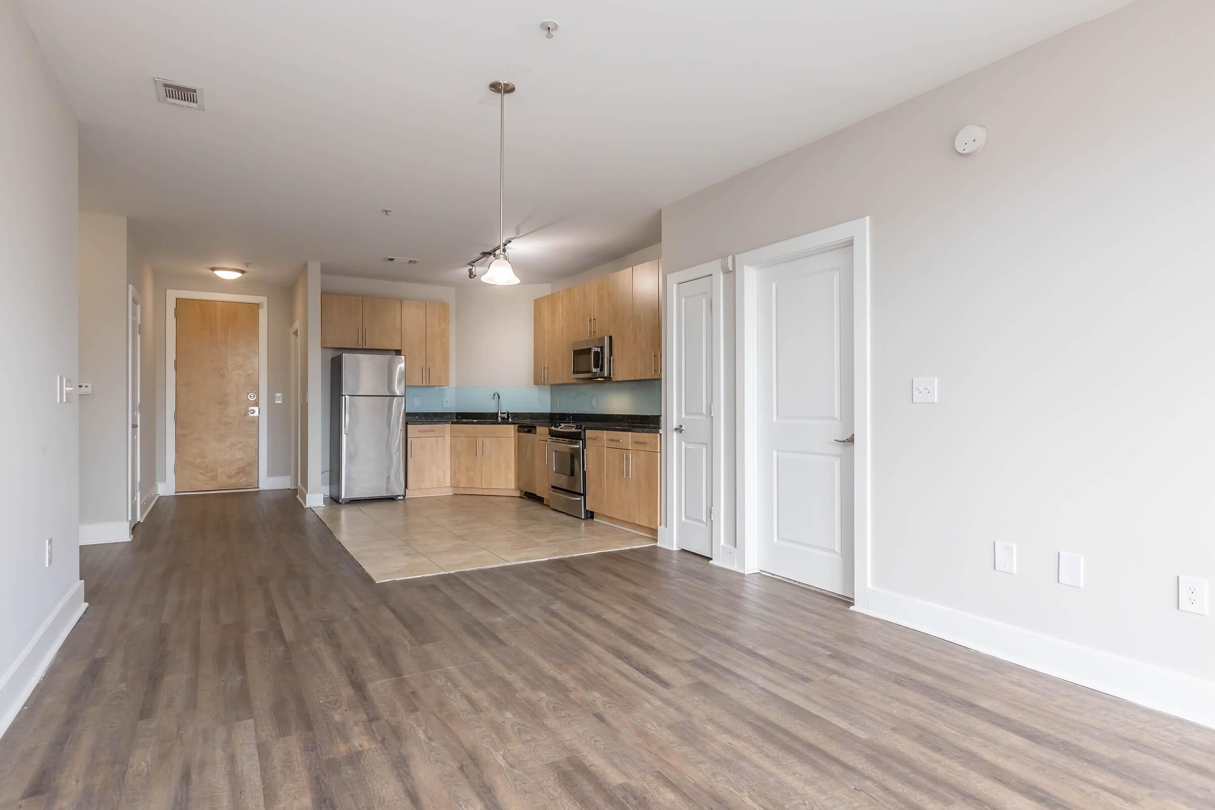 Spacious modern kitchen and living area with hardwood flooring, featuring wood cabinetry, stainless steel appliances, and a tile floor. Large windows provide natural light, and there’s a door leading to the entrance. Fresh, neutral wall colors create a bright and airy atmosphere.