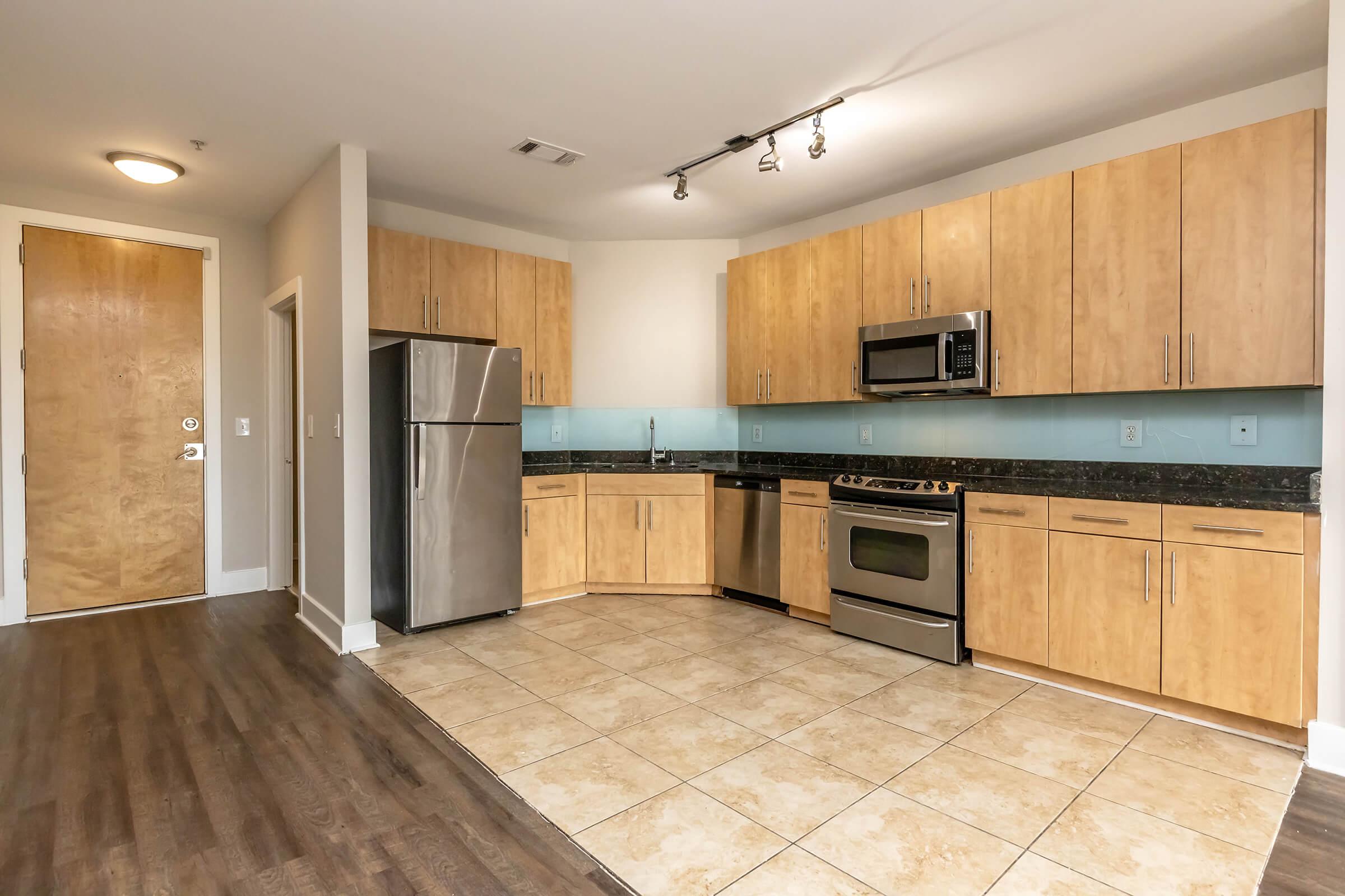 Modern kitchen featuring wooden cabinets, stainless steel refrigerator, microwave, and oven. The backsplash is a light blue tile, and the floor is a combination of tile and wood. A doorway is visible in the background, leading to an entrance. Overall, the space is bright and spacious with contemporary finishes.