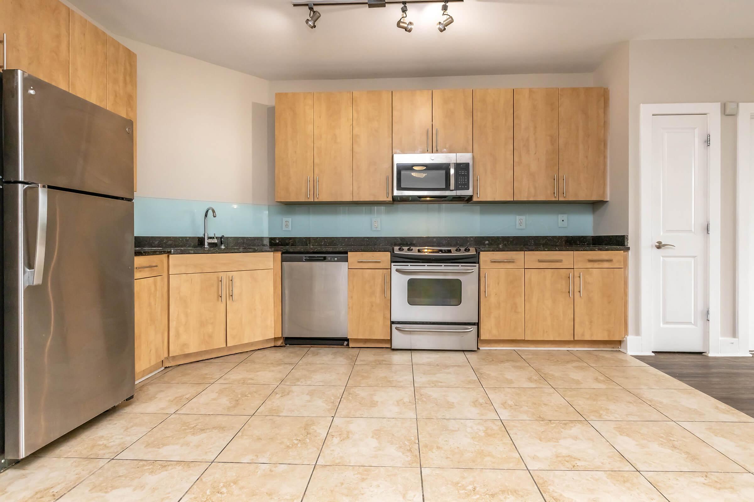 A modern kitchen featuring wooden cabinetry, stainless steel appliances including a refrigerator, microwave, and oven. The countertops are dark stone, and the backsplash is light blue glass. The floor is tiled with large beige tiles, and a door is visible on the right side.