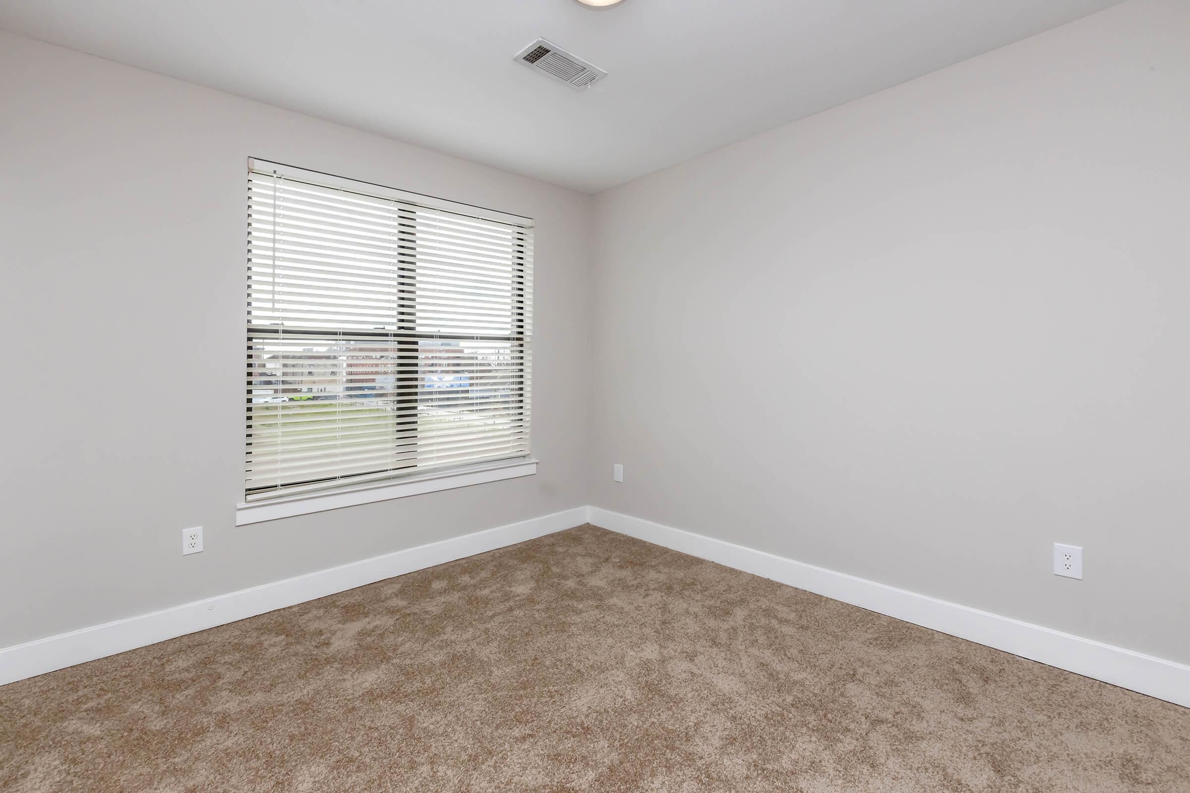 A spacious, empty bedroom featuring light beige carpet and neutral-colored walls. A large window with white blinds allows natural light to illuminate the room. The window looks out onto a view with buildings in the distance, providing an airy and bright atmosphere.