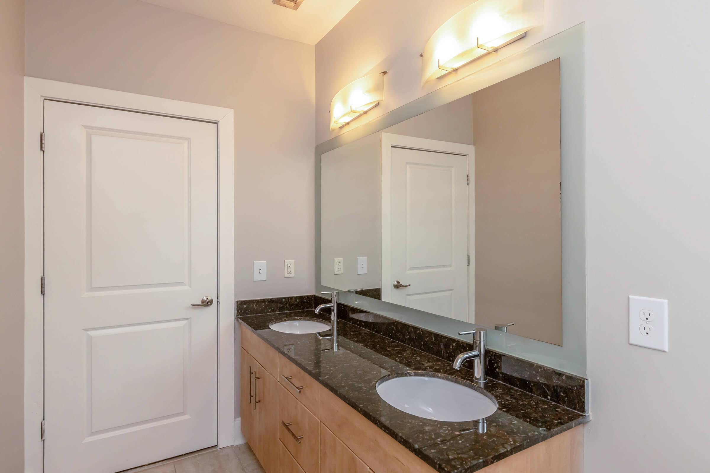 A modern bathroom featuring a double vanity with two circular sinks, a large mirror above, and stylish wall-mounted lights. The cabinetry is light wood, and the countertop is dark with a granite finish. A white door leads to another room, with neutral-colored walls and tile flooring.