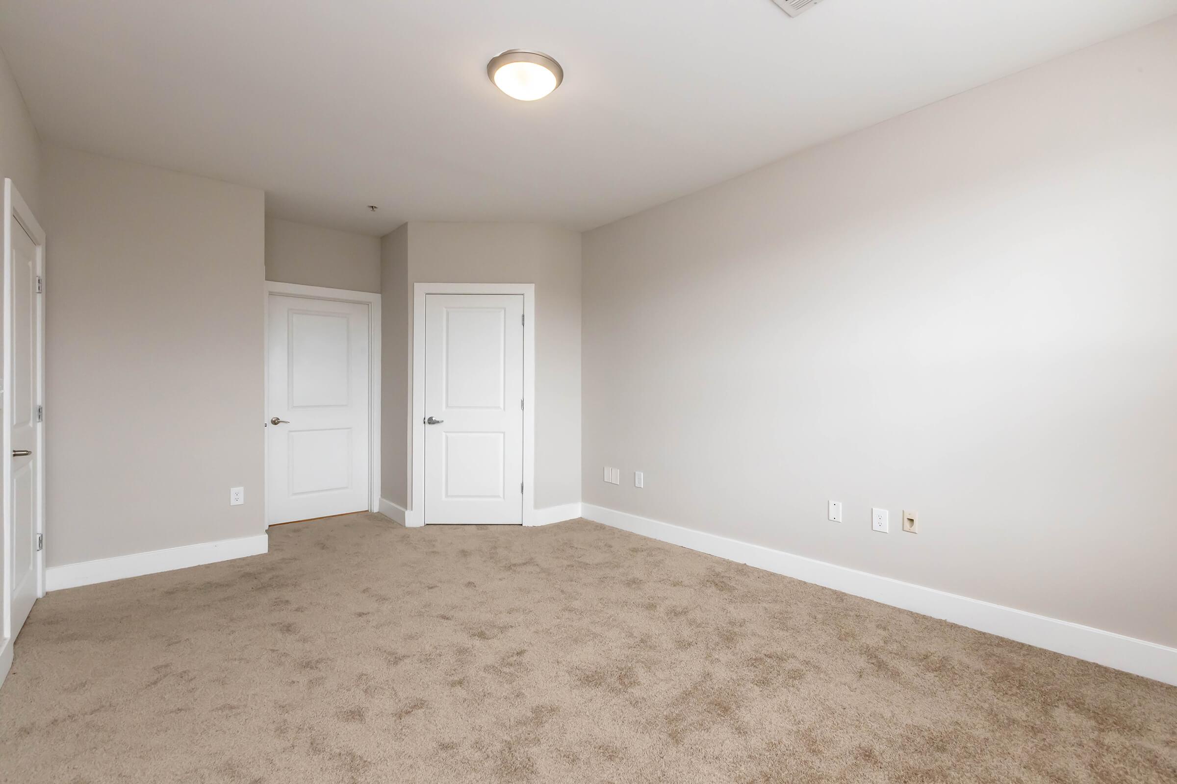 A spacious, empty room with beige carpet and light gray walls. It features two white doors, one closed and one open, leading to an adjacent space. The ceiling has a simple light fixture providing overhead illumination, creating a bright and airy ambiance.