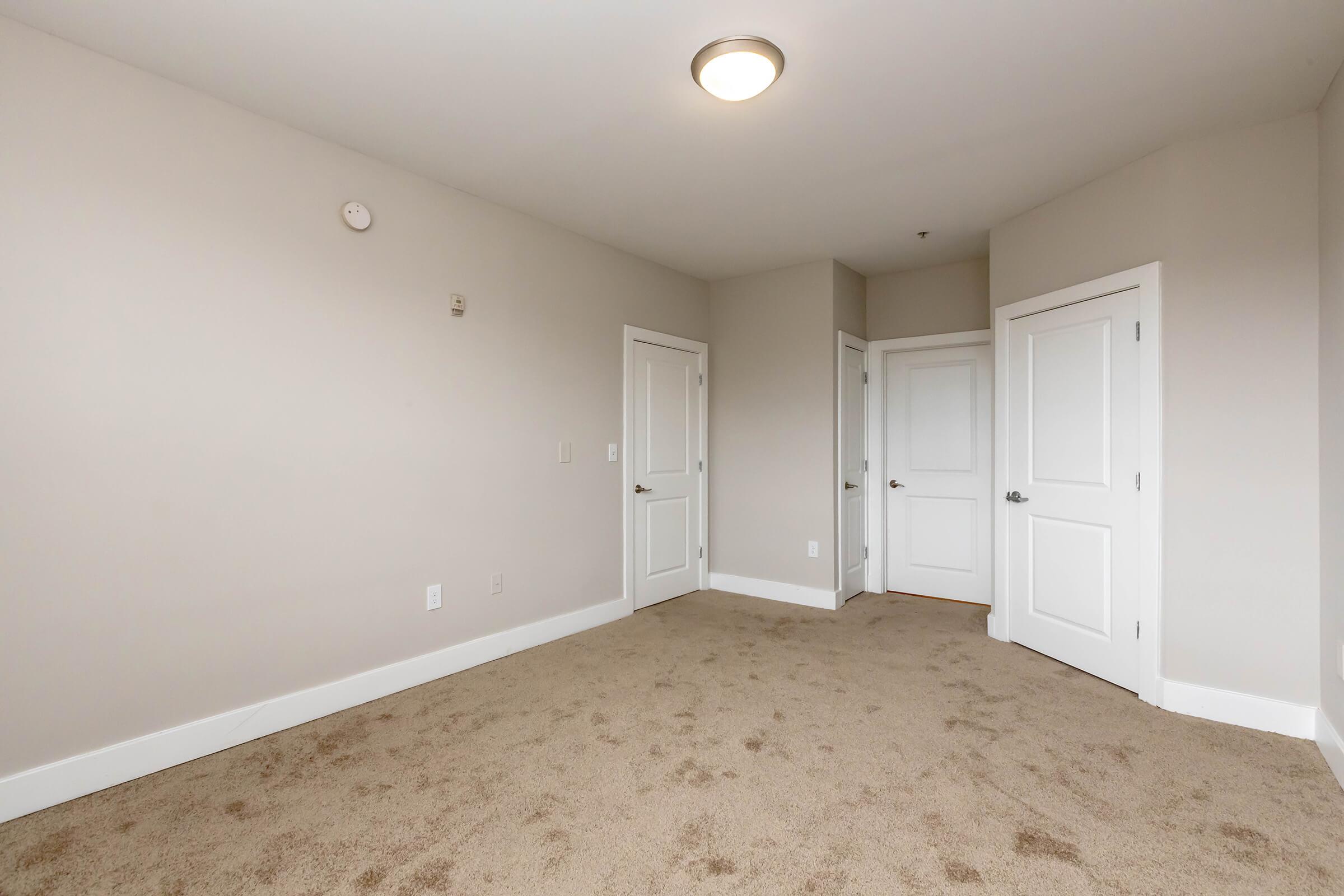 A spacious, empty room with light beige walls and plush, light brown carpet. Two closed doors are visible on the right side, and a small round ceiling light provides overhead illumination. The room has a neutral and airy feel, making it suitable for various uses.
