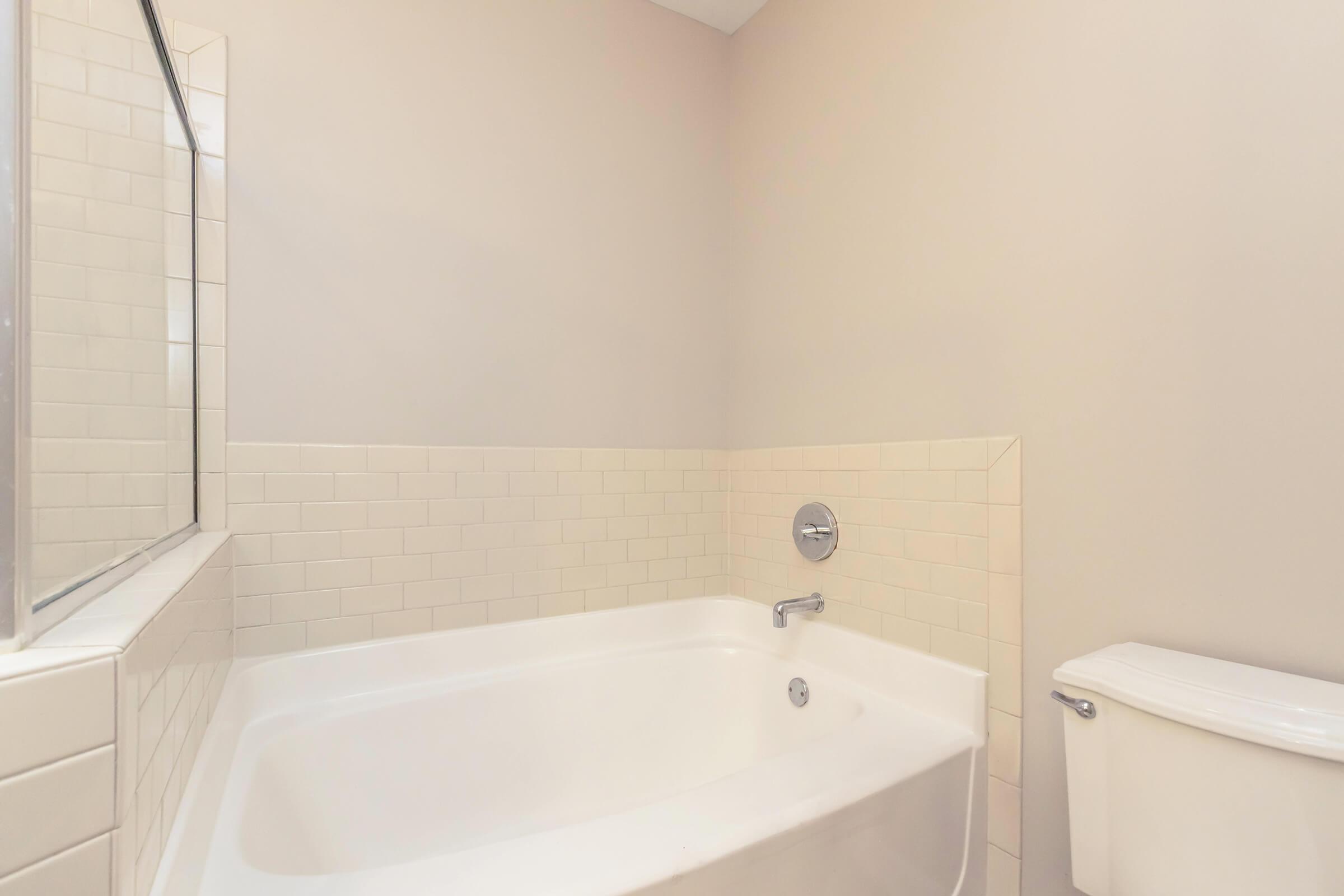 A clean, modern bathroom featuring a white bathtub with chrome fixtures, a wall-mounted mirror, and a white toilet. The walls are light-colored, and the bathtub is surrounded by beige tiled accents, giving the space a minimalist and airy feel.