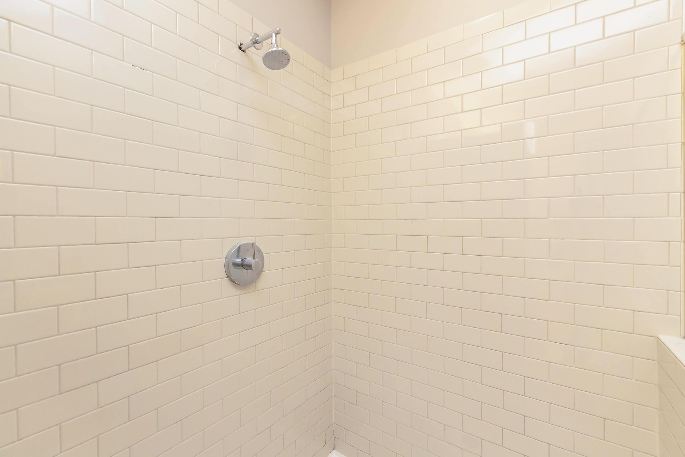 A clean, modern shower with light-colored subway tile walls. The shower features a chrome showerhead and a water control knob, set against a bright, minimalistic background. The floor is not visible, and the overall design emphasizes simplicity and functionality.