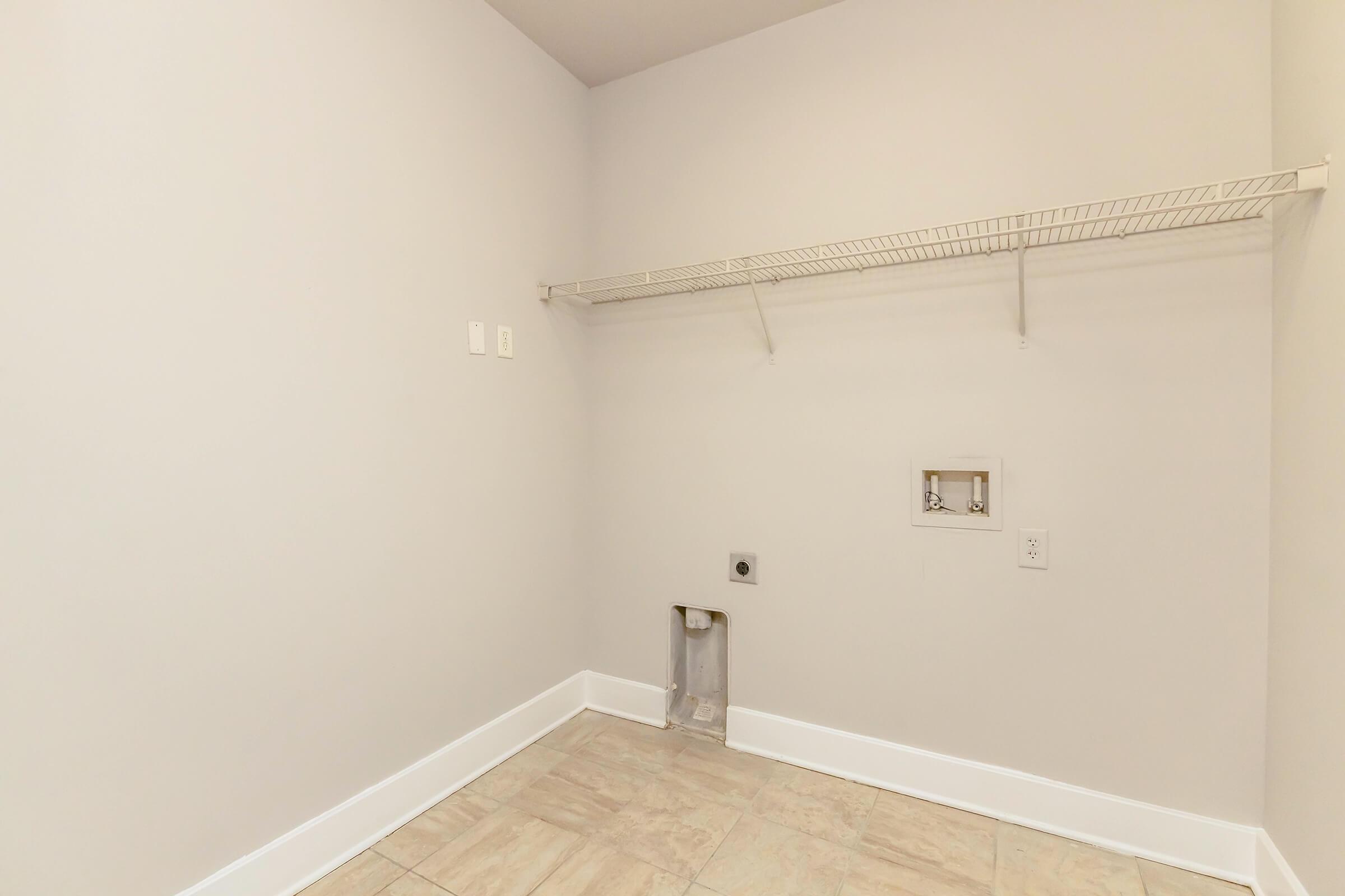 Empty laundry room with gray walls and a tiled floor. A metal wire shelf is attached to the wall, and there are two electrical outlets. A plumbing connection is visible on the floor, indicating a space for a washing machine. The room is well-lit and tidy, emphasizing a minimalist design.