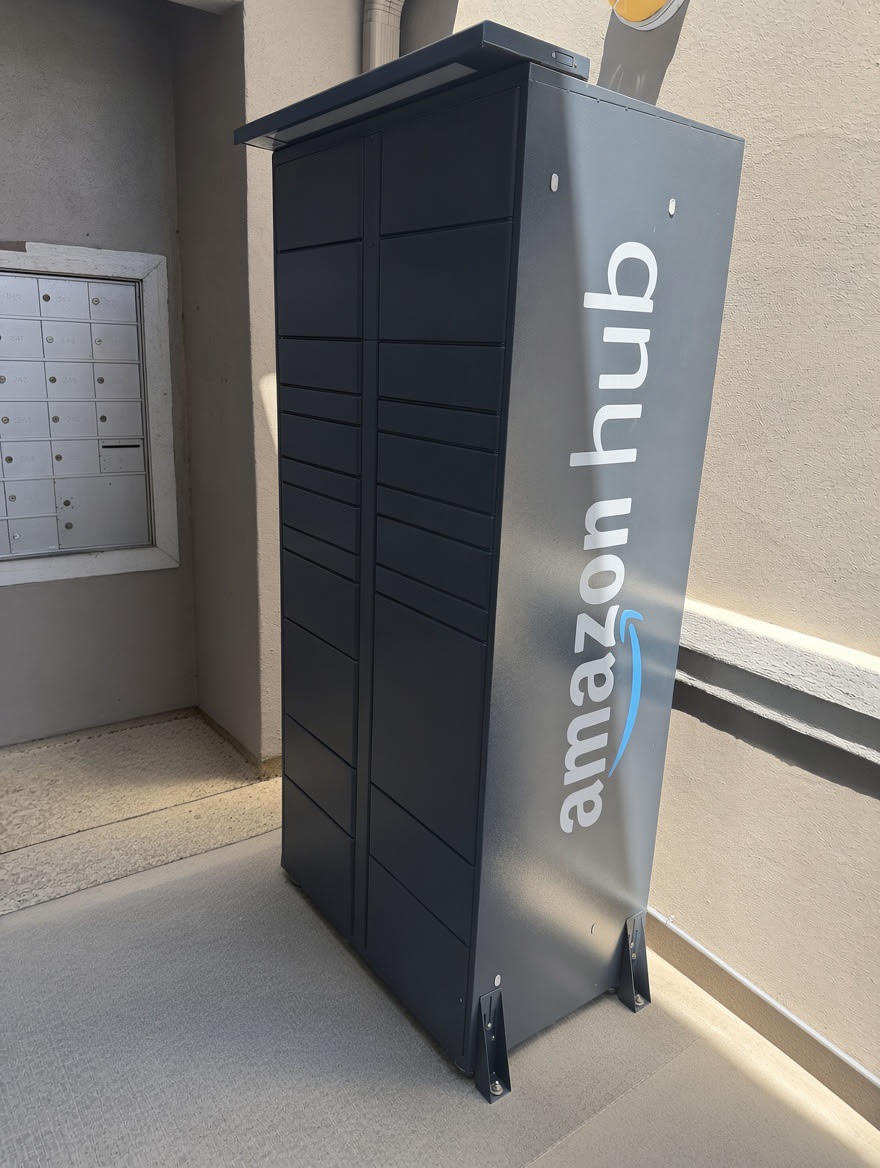 A gray Amazon Hub locker positioned in a hallway. The locker has the Amazon logo and offers a secure location for package pickups. Nearby, there are mailboxes, suggesting it's in an apartment or residential complex. The floor is concrete, and the environment appears well-lit.