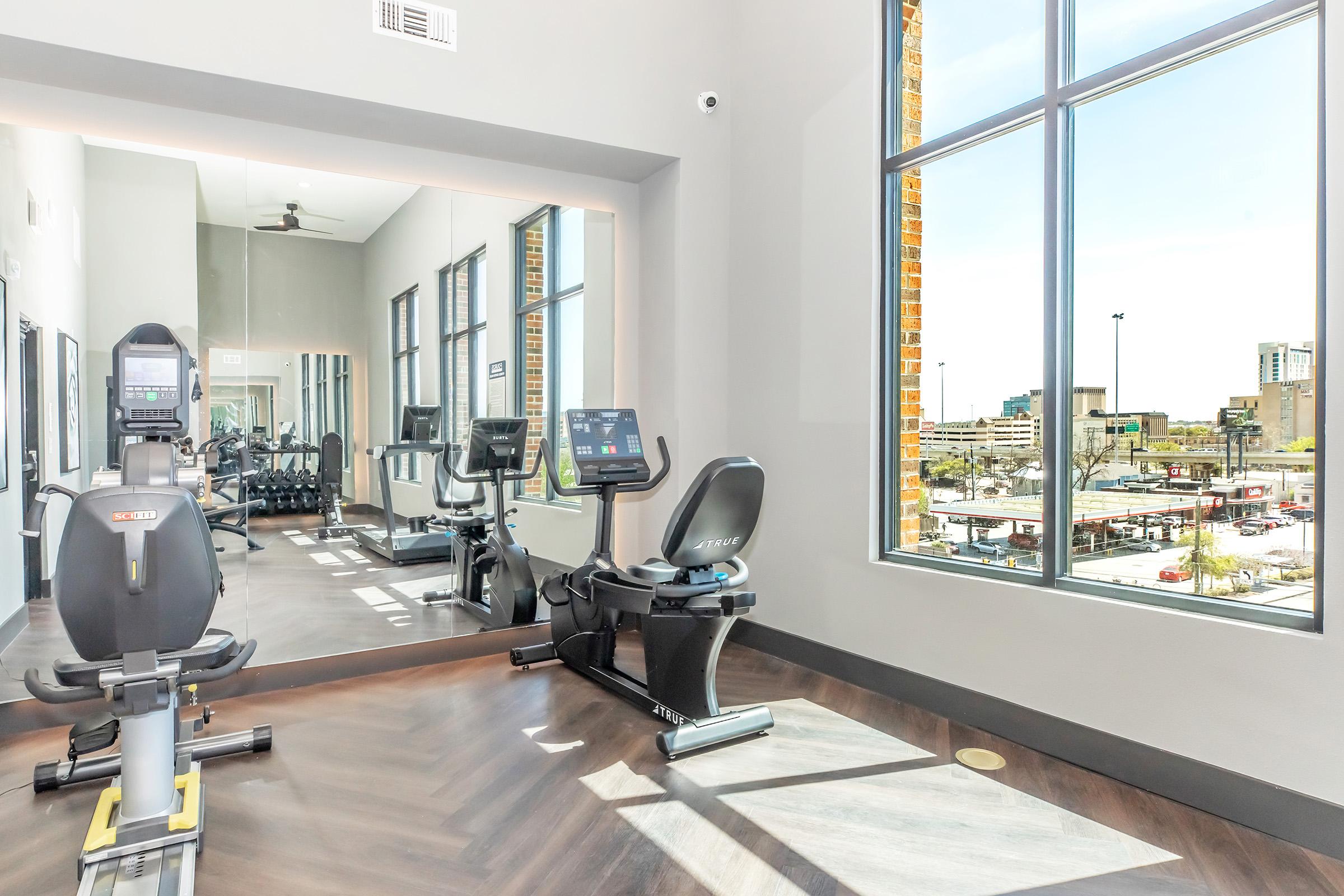 A bright, modern fitness room featuring exercise equipment, including stationary bikes and a large mirror. Natural light streams through large windows, offering a view of an outdoor area and surrounding cityscape. The space has a contemporary design with wooden flooring and neutral wall colors.
