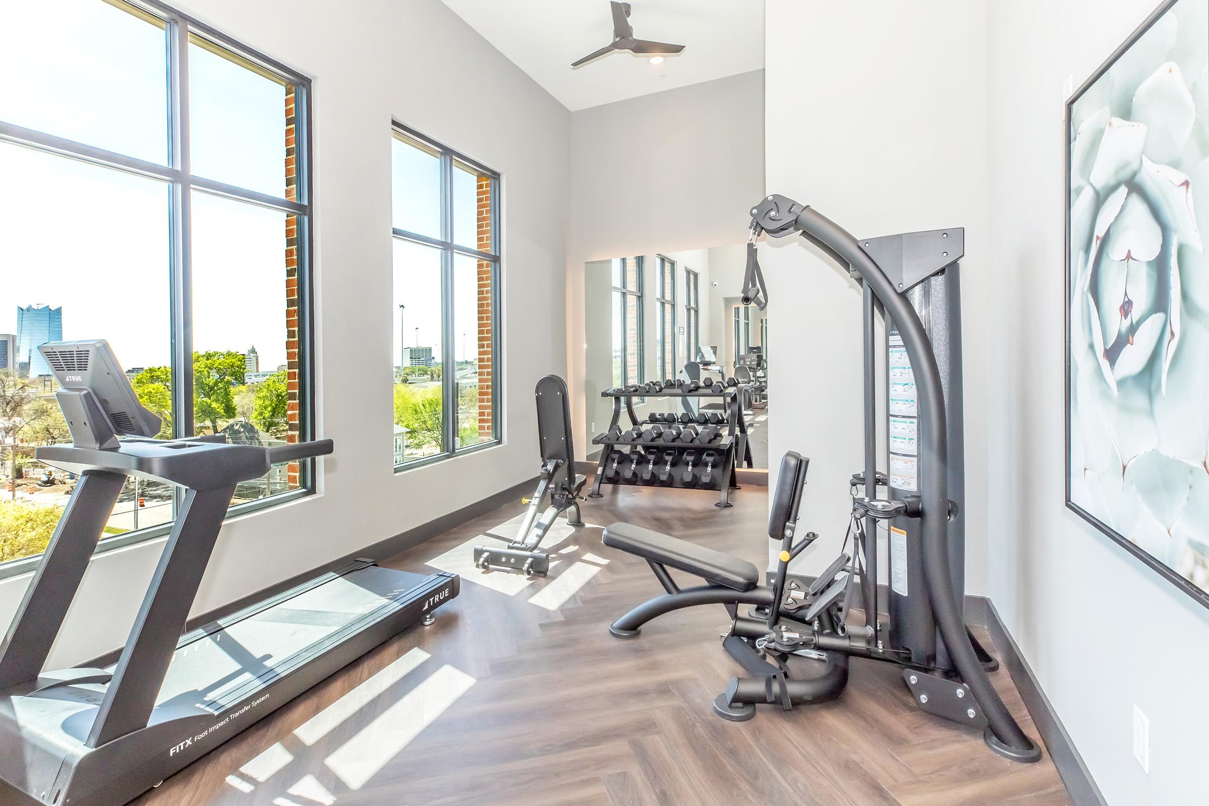 A modern gym interior featuring a treadmill, weightlifting equipment, and a multi-station machine. Large windows provide natural light and a view of the outdoors. The flooring is wood-like, and the walls are painted in neutral tones, creating a bright and inviting workout space.