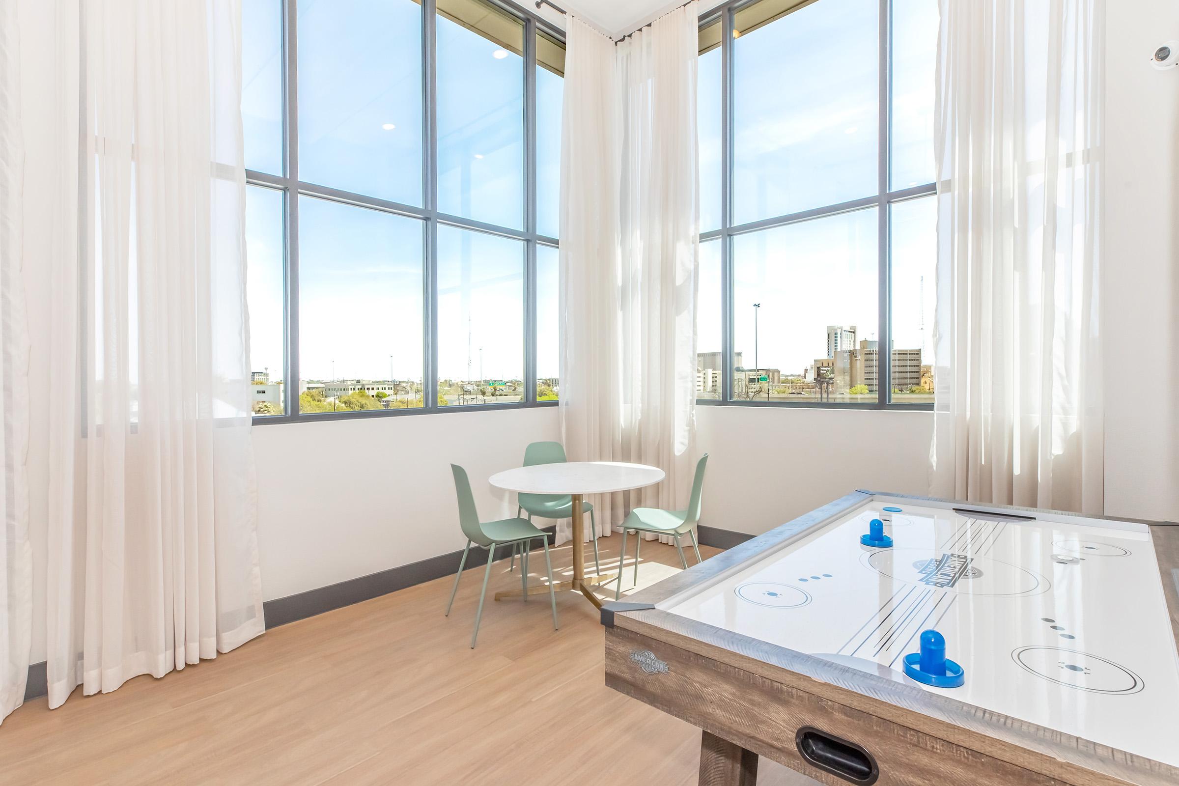 Bright and airy room featuring large windows with sheer curtains, a small round table with two green chairs, and a tabletop air hockey game. The view outside includes a cityscape. The space emphasizes a modern and welcoming atmosphere.