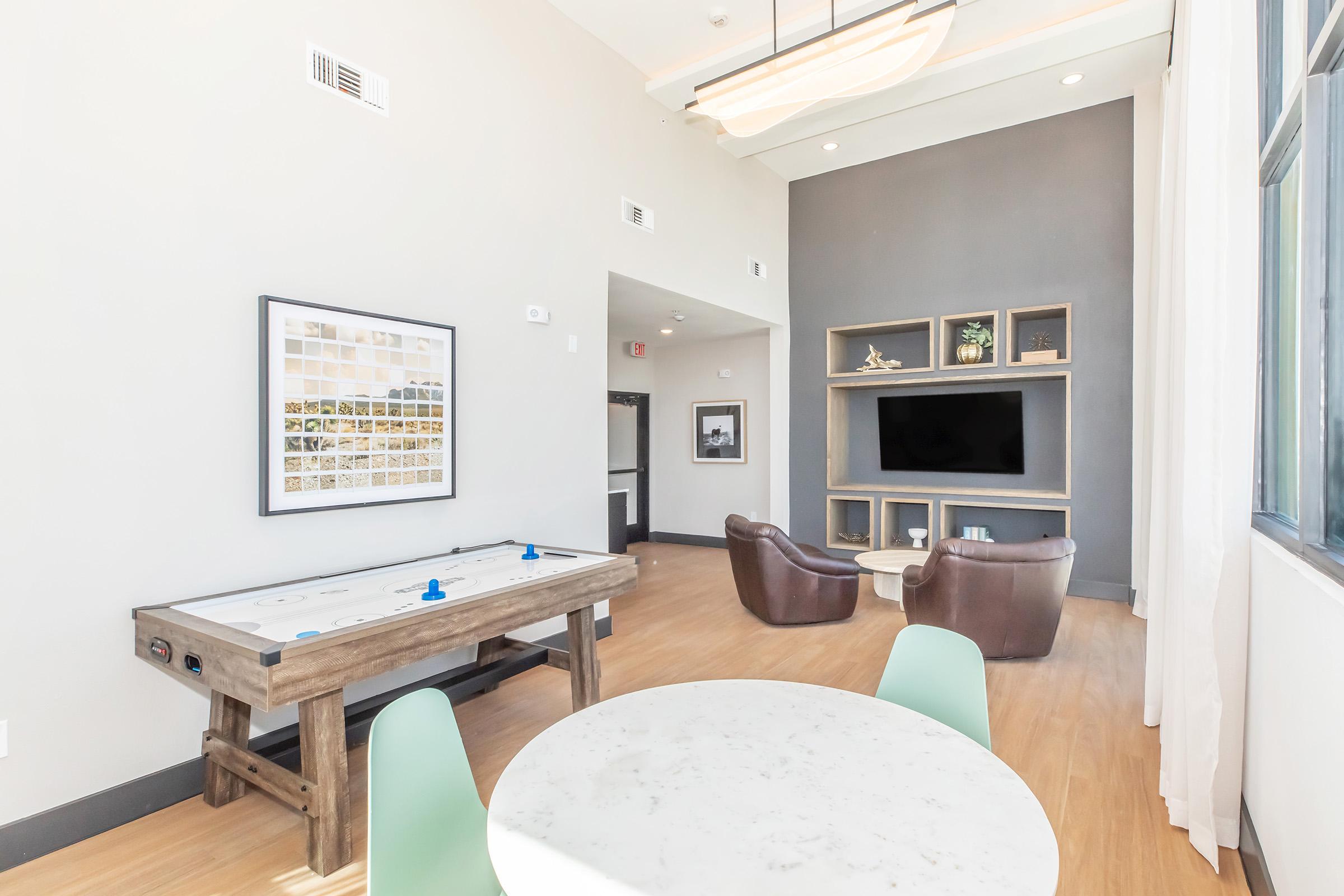 A modern indoor lounge featuring a marble-topped table with mint green chairs, a wooden air hockey table, two brown leather armchairs, and a wall-mounted TV. Soft natural light enters through large windows, accentuating the neutral-colored walls and contemporary decor.