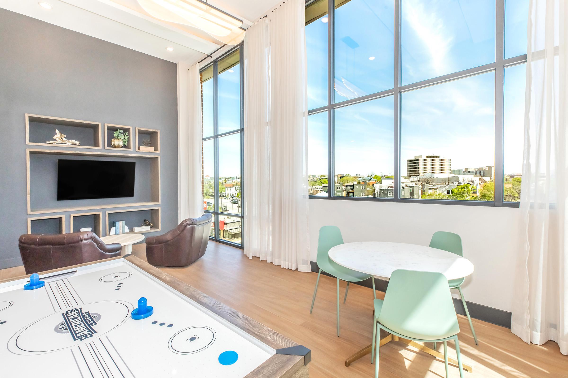 Bright room with large windows showcasing an urban view. A modern air hockey table is in the foreground, with blue pucks and a scoreboard. There are two comfortable brown armchairs and a small circular table with light green chairs nearby. The space has a light, airy feel, enhanced by ample natural light.