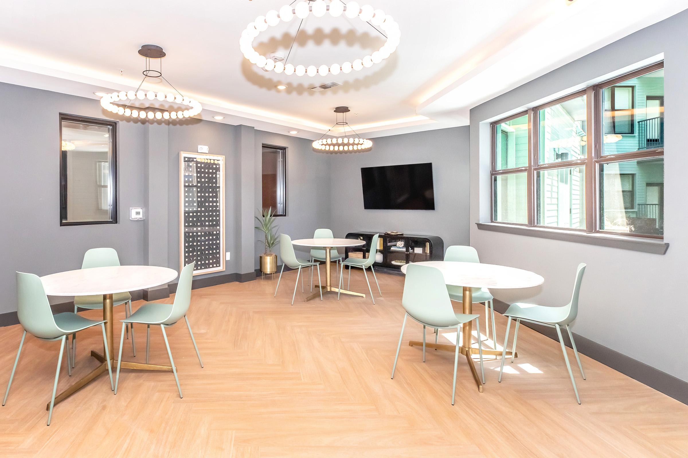A modern common area featuring two round tables with mint green chairs, light wooden flooring, and large windows. There are two elegant chandelier light fixtures hanging from the ceiling and a flat-screen TV mounted on the wall. A decorative plant is placed near a corner.