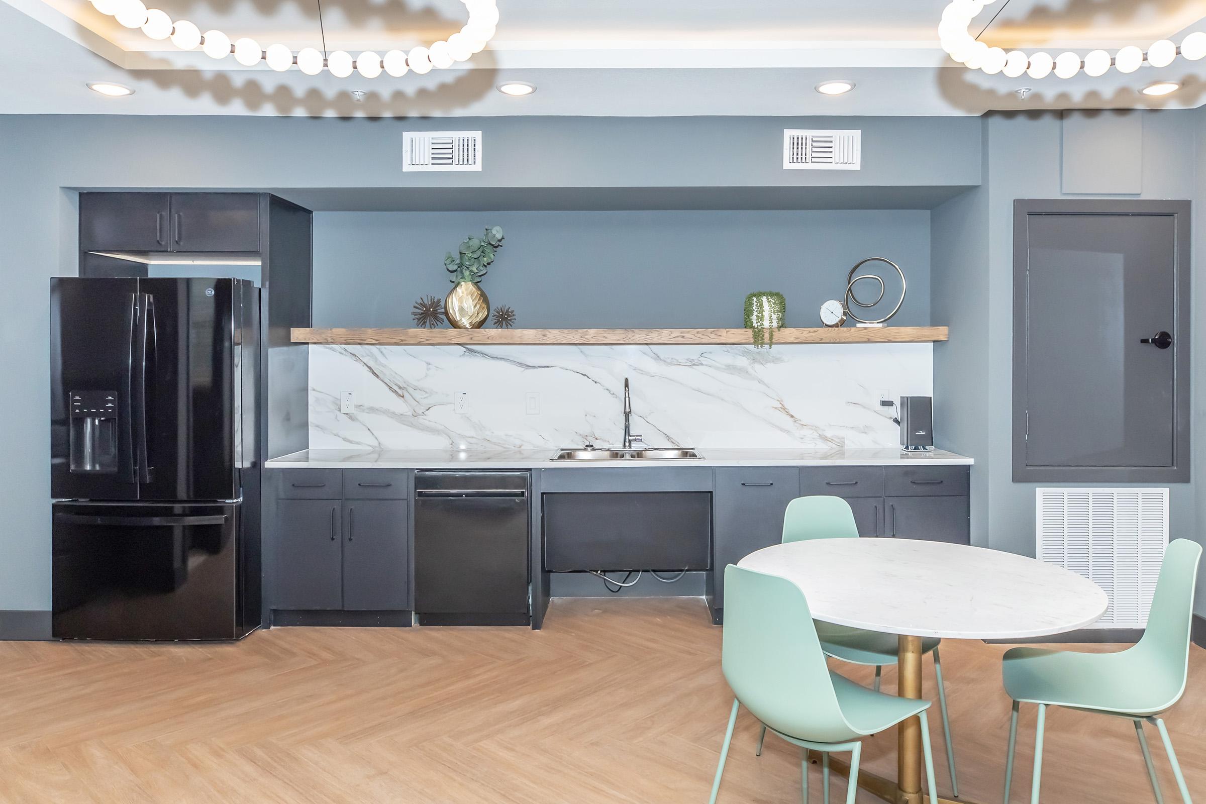 Modern kitchen featuring dark cabinets, a marble backsplash, and stainless steel appliances. A round dining table with light green chairs is in the foreground. The room is well-lit with stylish circular light fixtures.