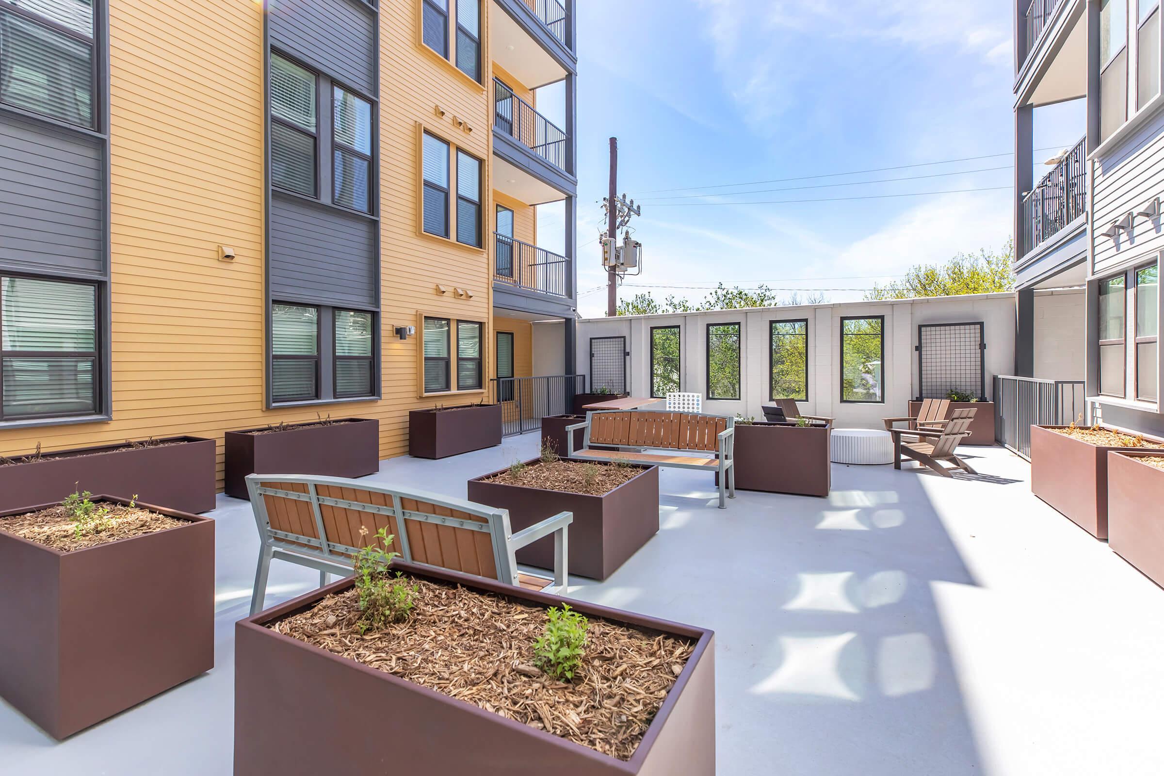 An urban courtyard featuring modern architecture with yellow and gray exterior walls, surrounded by planters with greenery. There are benches for seating and a high pole, possibly for cellular service, in the background. The area is brightly lit with blue skies visible.