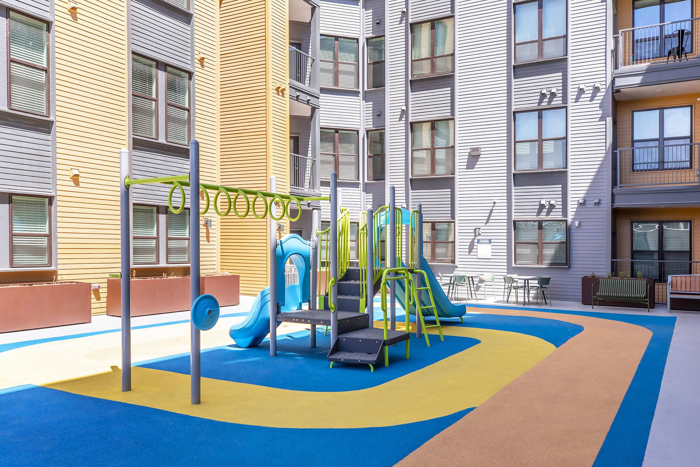 Colorful playground with slides, climbing structures, and rings, surrounded by modern apartment buildings. The ground features soft, blue and brown mats, providing a safe play area. Benches and tables are visible in the background, offering space for relaxation.