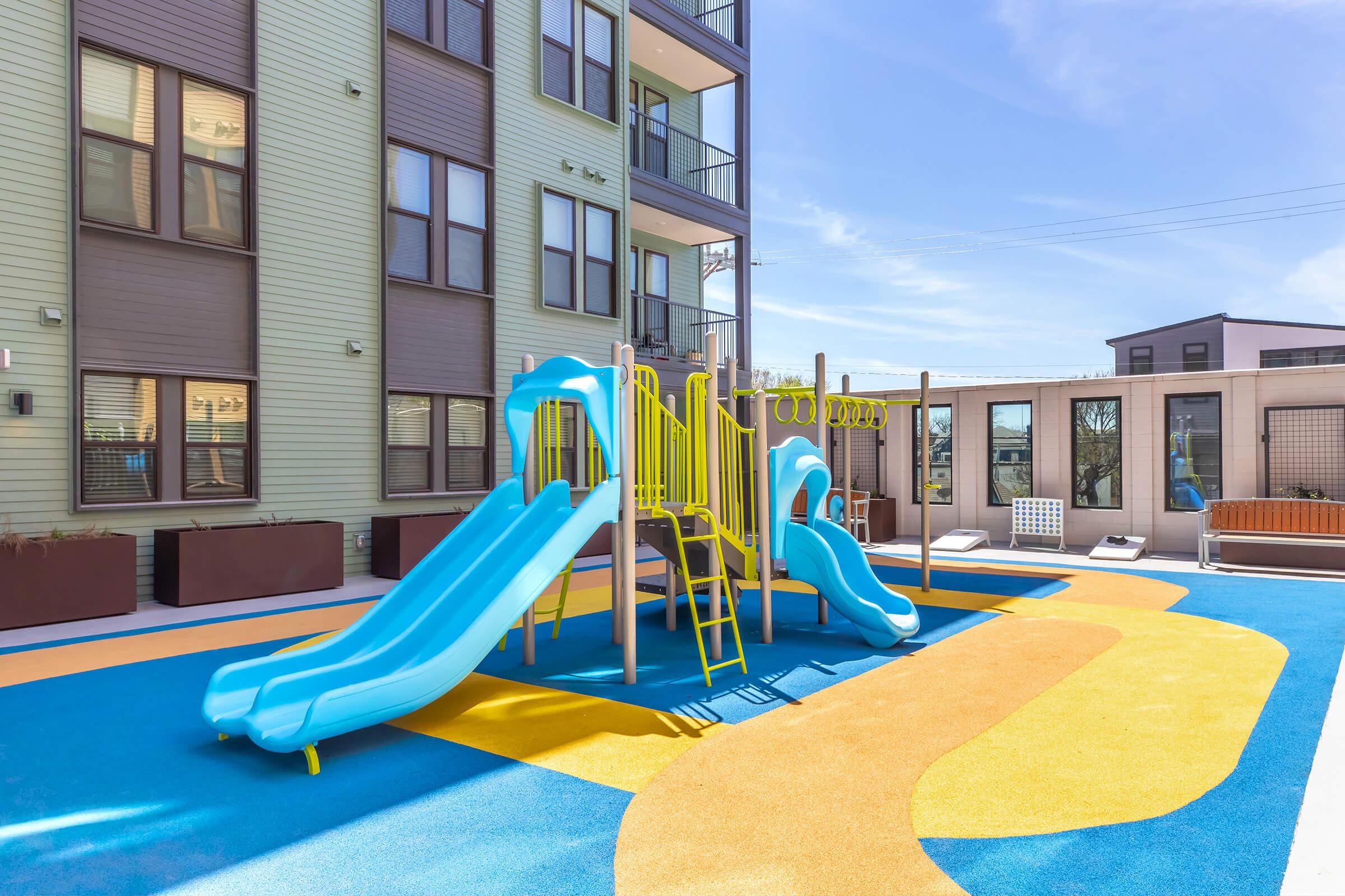 Playground area featuring colorful blue slides, surrounded by bright yellow and blue textured flooring. In the background, modern apartment buildings are visible, with a fenced area and outdoor seating. The sky is clear, indicating a sunny day.