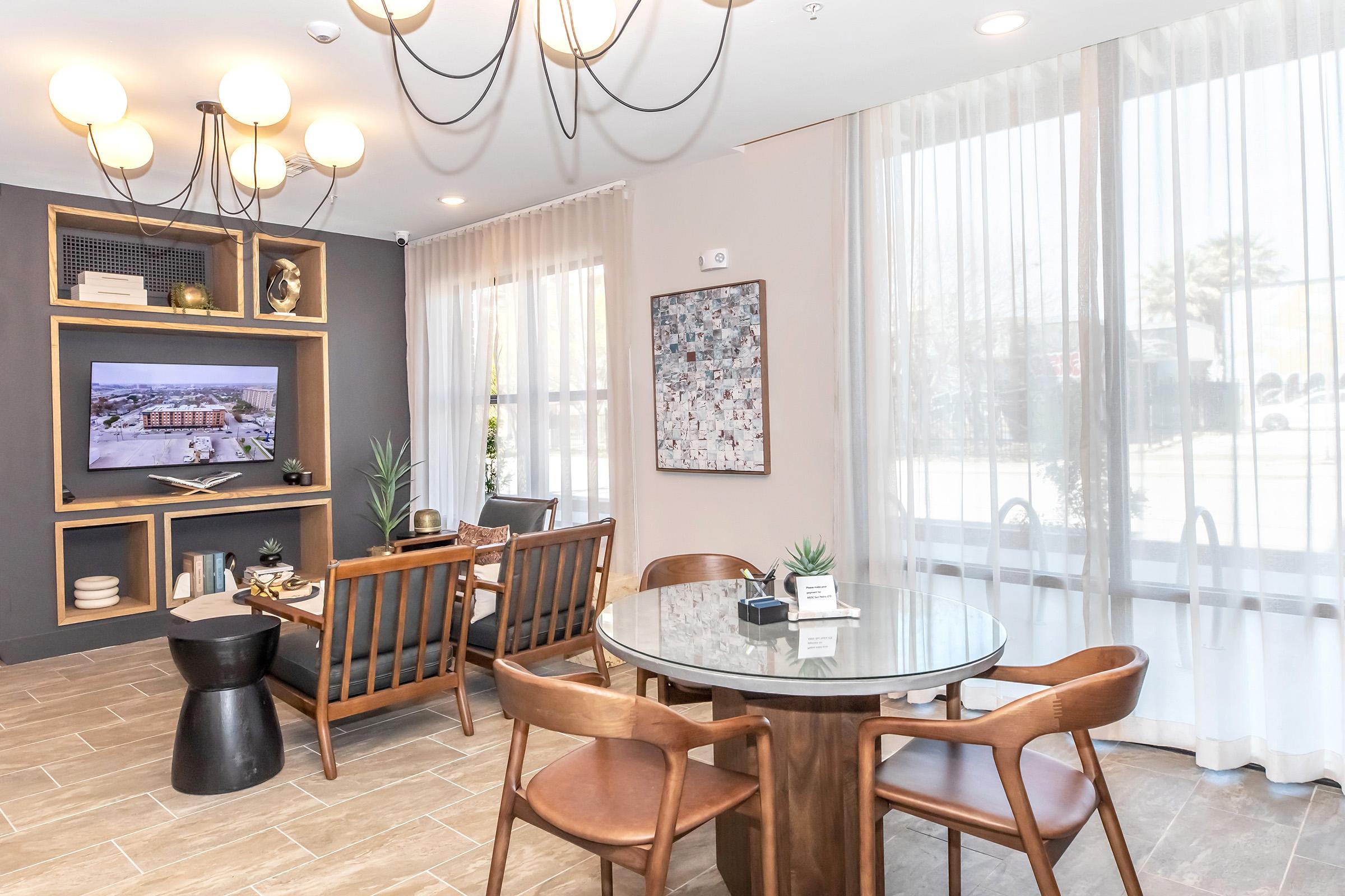 A modern lounge area featuring a round table with wooden chairs, a black side table, and a large TV mounted on a wall. Decor includes a contemporary chandelier, plants, and a gallery of framed images. The space has large windows with sheer curtains, allowing natural light to fill the room.