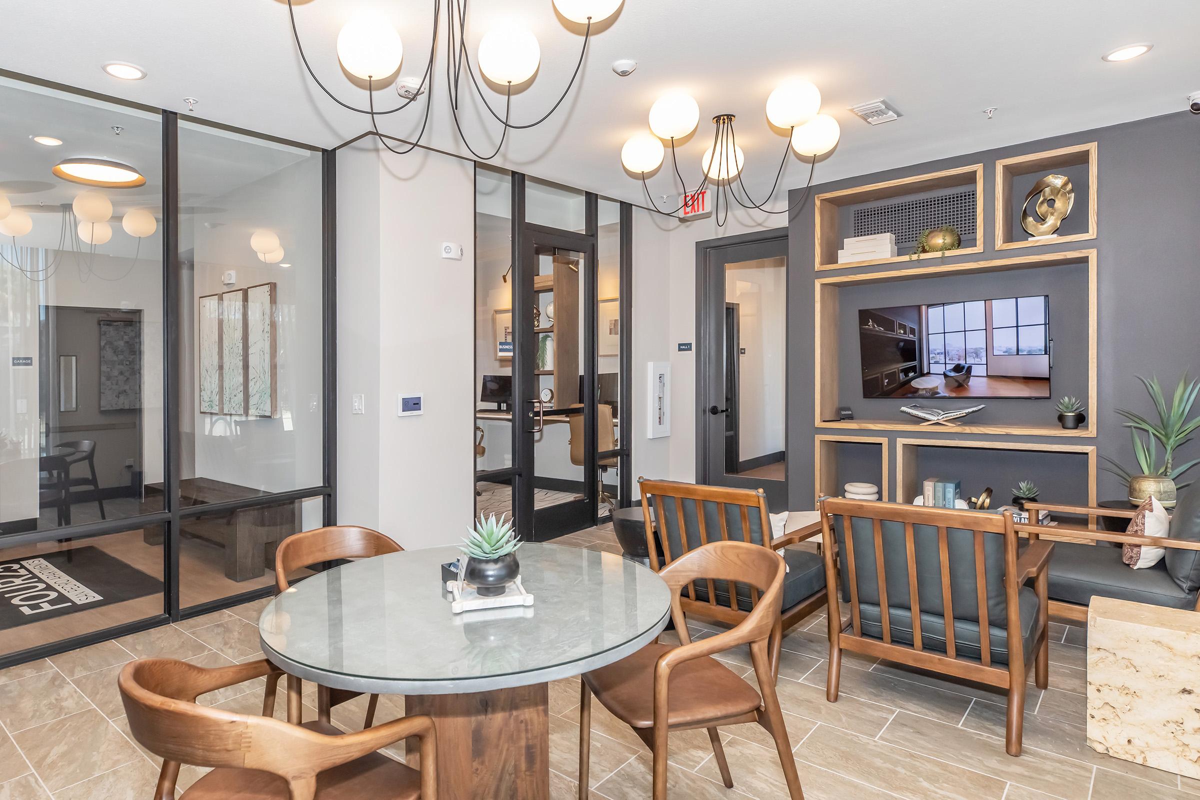 A modern lounge area featuring a round glass table surrounded by wooden chairs. The space is well-lit with stylish pendant lighting. A decorative wall unit with a TV, plants, and art frames adds visual interest. Large windows allow natural light, creating an inviting atmosphere.