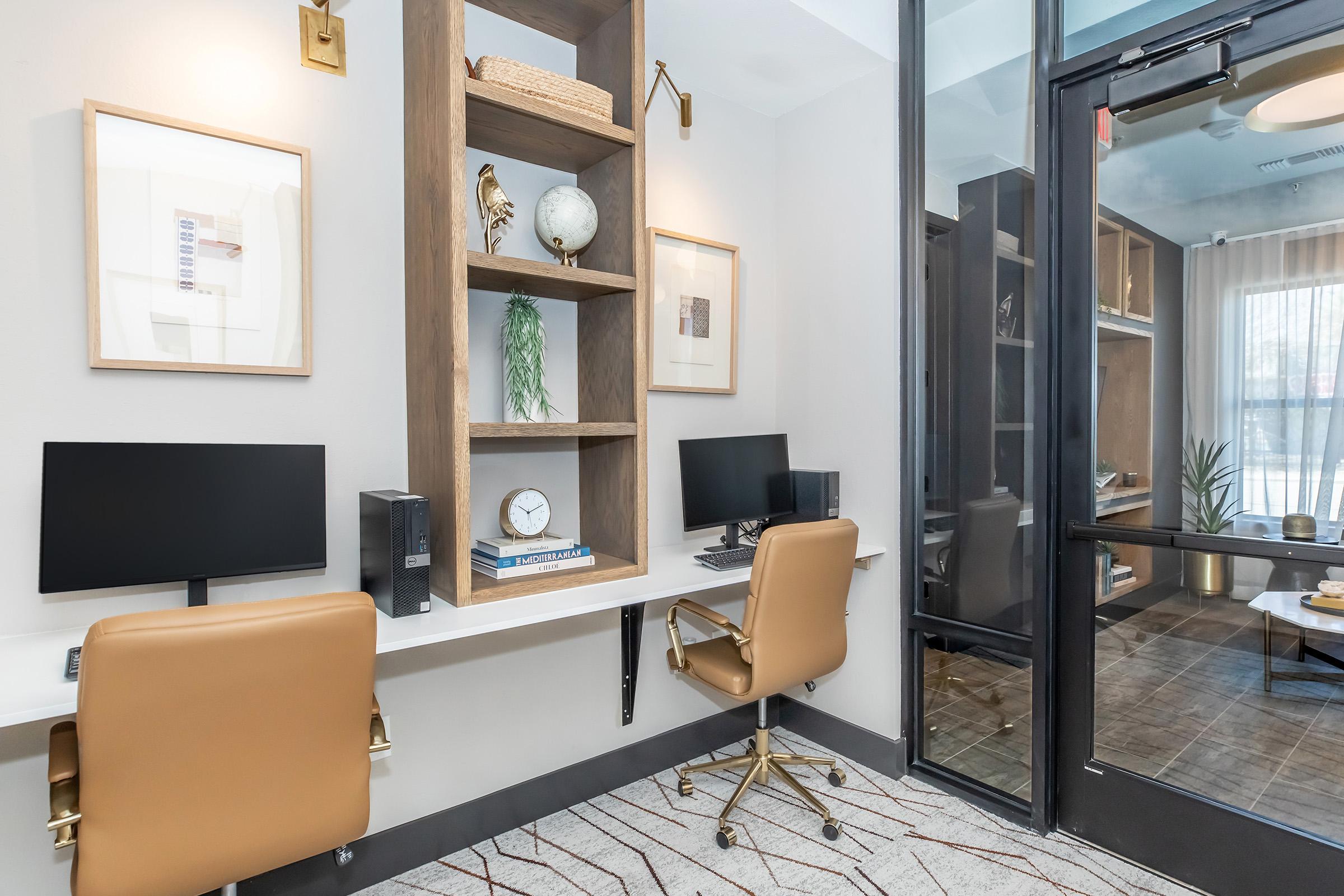 A modern office space featuring two desk setups, each with a computer monitor and a stylish brown chair. The walls are adorned with framed art and decorative items on shelves. A glass door leads to another room, and natural light streams in through a nearby window. The overall design is sleek and contemporary.