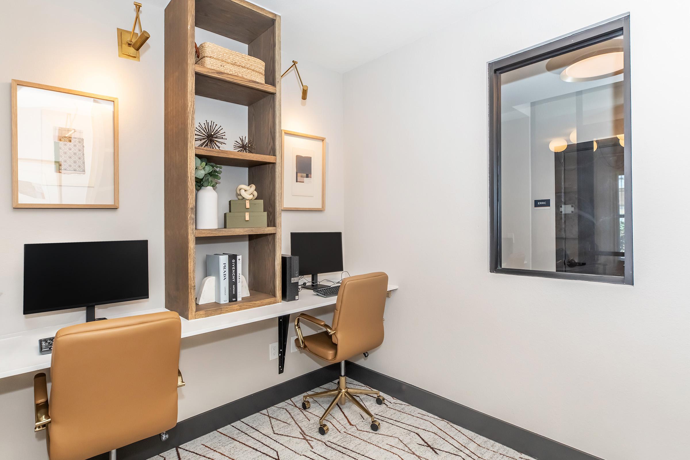 Two modern workstations with sleek black monitors and stylish tan chairs are arranged in a minimalist office space. A decorative wooden shelf is mounted on the wall, featuring framed art, a woven basket, and decorative objects. The room has a neutral color palette and contemporary lighting.