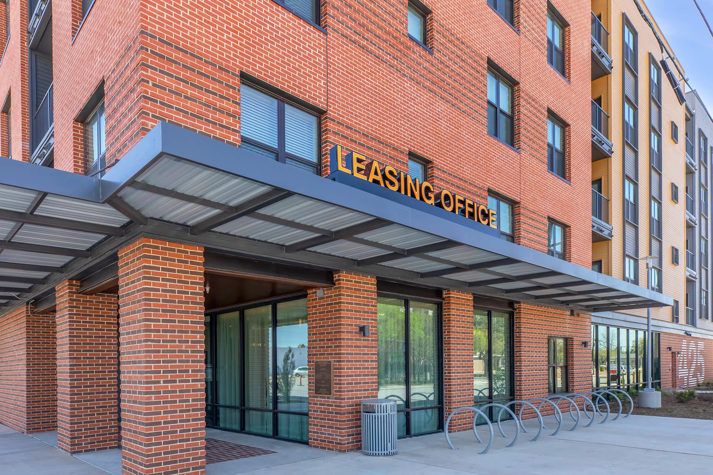 A modern leasing office with a brick exterior and large windows, featuring a covered entrance. The sign above the entrance reads "LEASING OFFICE." Bicycles are parked nearby, and there are landscaped areas visible. The building has multiple stories with a contemporary design.
