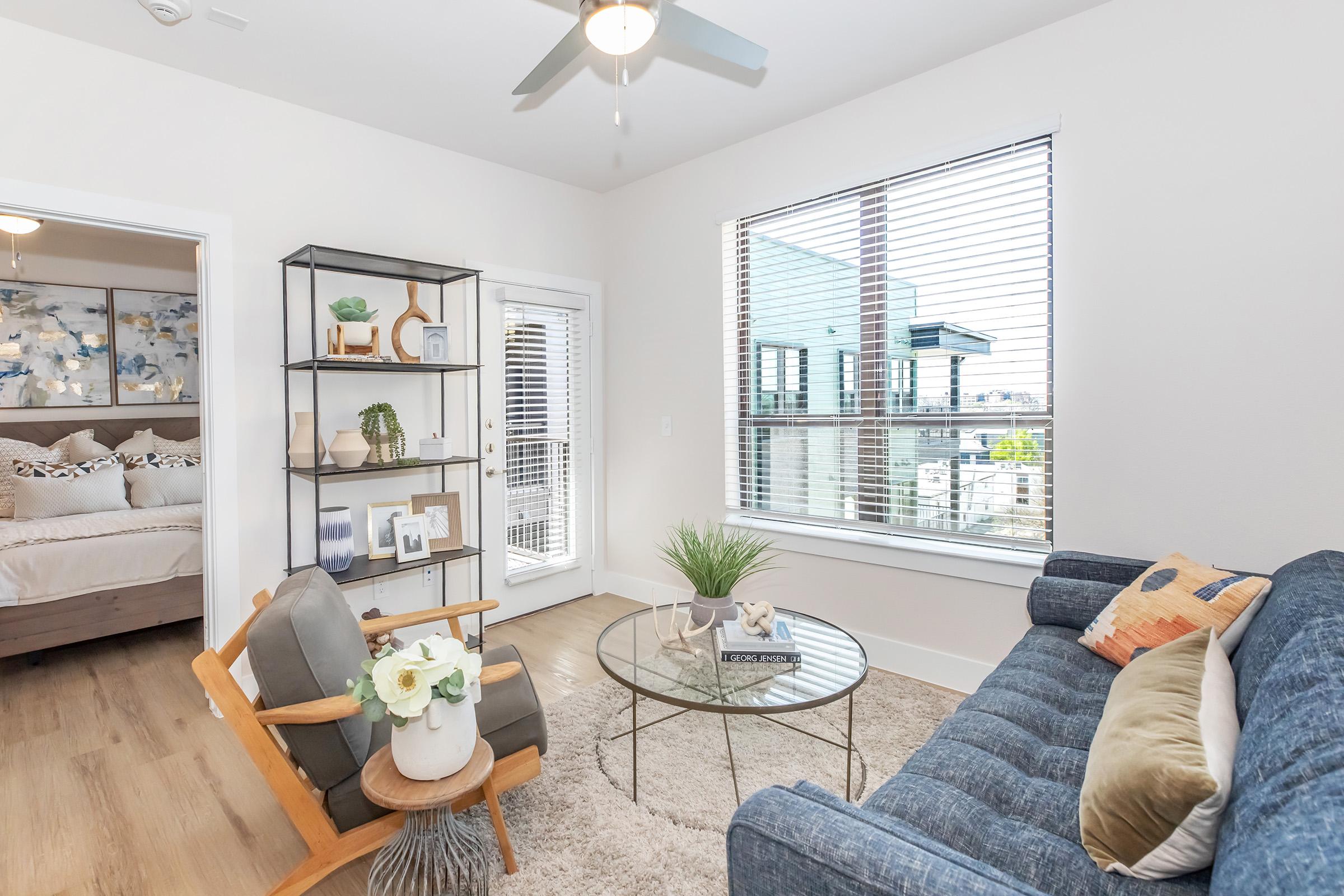 A cozy living room featuring a blue sofa, a glass coffee table with a plant and decor, and a wooden armchair. A bookshelf displays plants and framed photos. Natural light streams through large windows with blinds, revealing a view of an outdoor balcony. A bedroom is visible in the background.