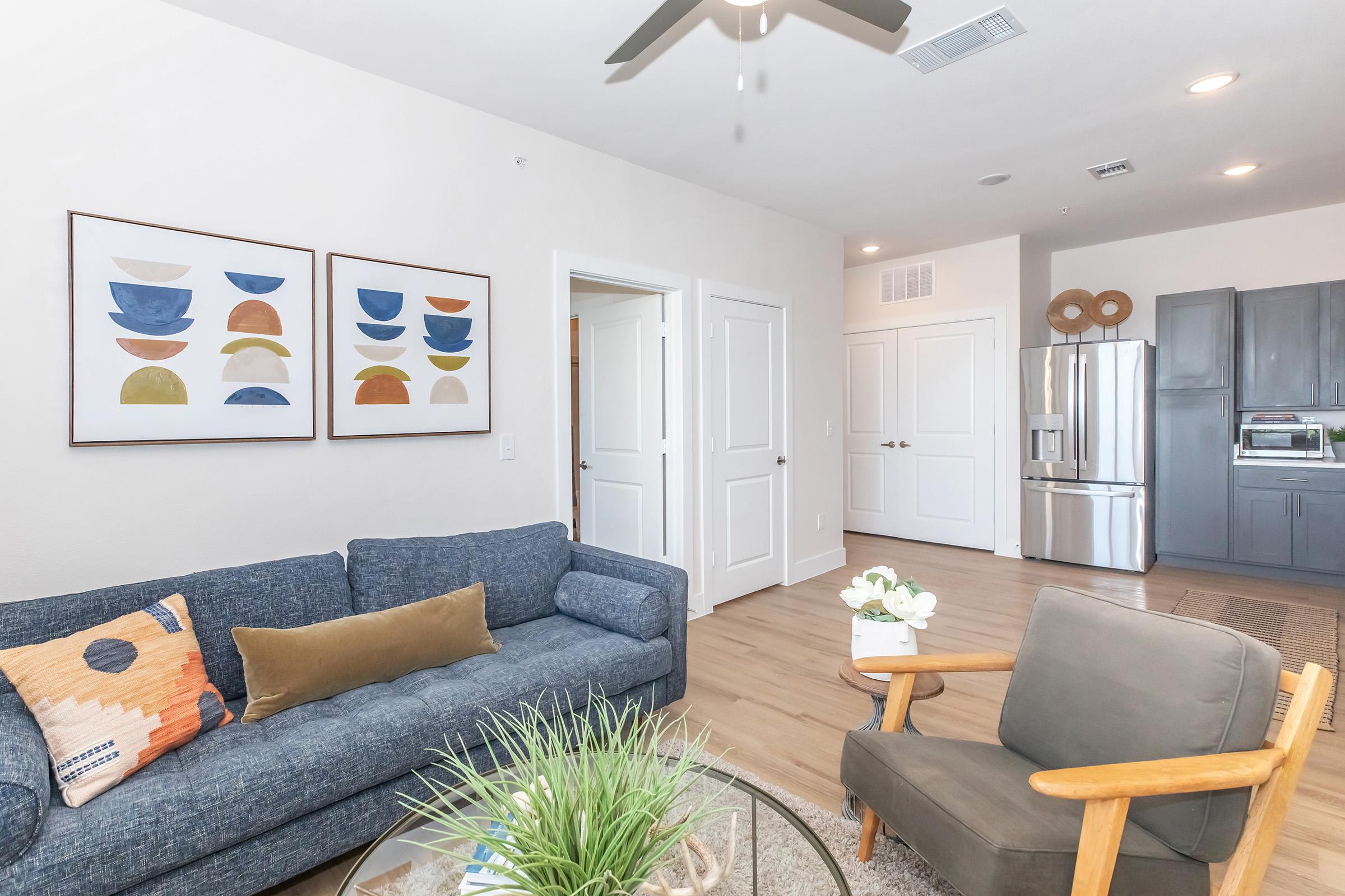 A bright and modern living room featuring a blue sofa with decorative pillows and a wooden chair. Two abstract framed artworks are displayed on the wall, and a small table holds a plant and a vase. A kitchen area with stainless steel appliances is visible in the background. Natural light fills the space.