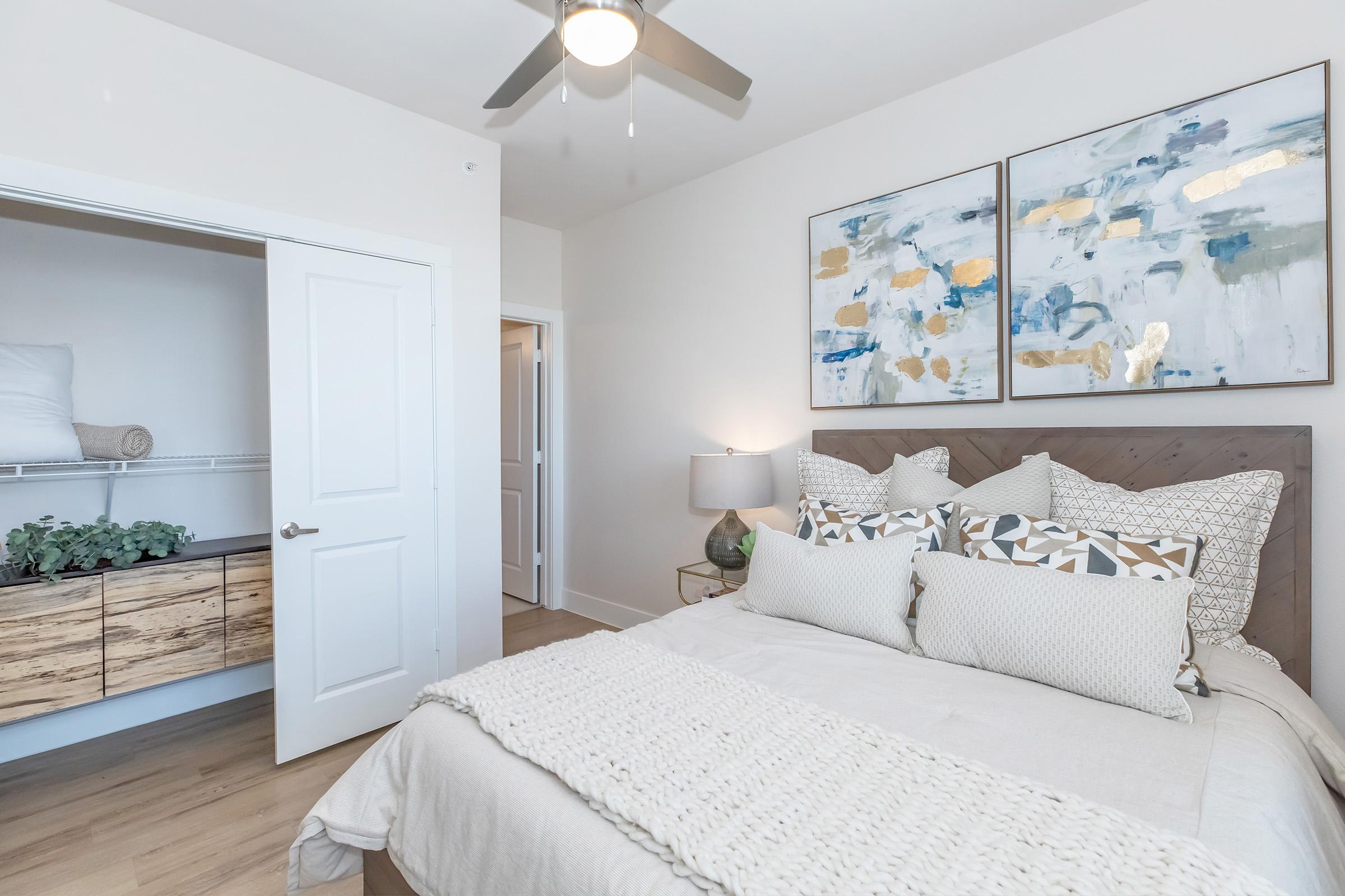 A stylish bedroom featuring a comfortable bed with decorative pillows, a light blanket, and a wooden headboard. The walls are adorned with abstract paintings. A dresser with a minimalist design is visible, alongside an open closet door. Natural light filters in, creating a warm and inviting atmosphere.