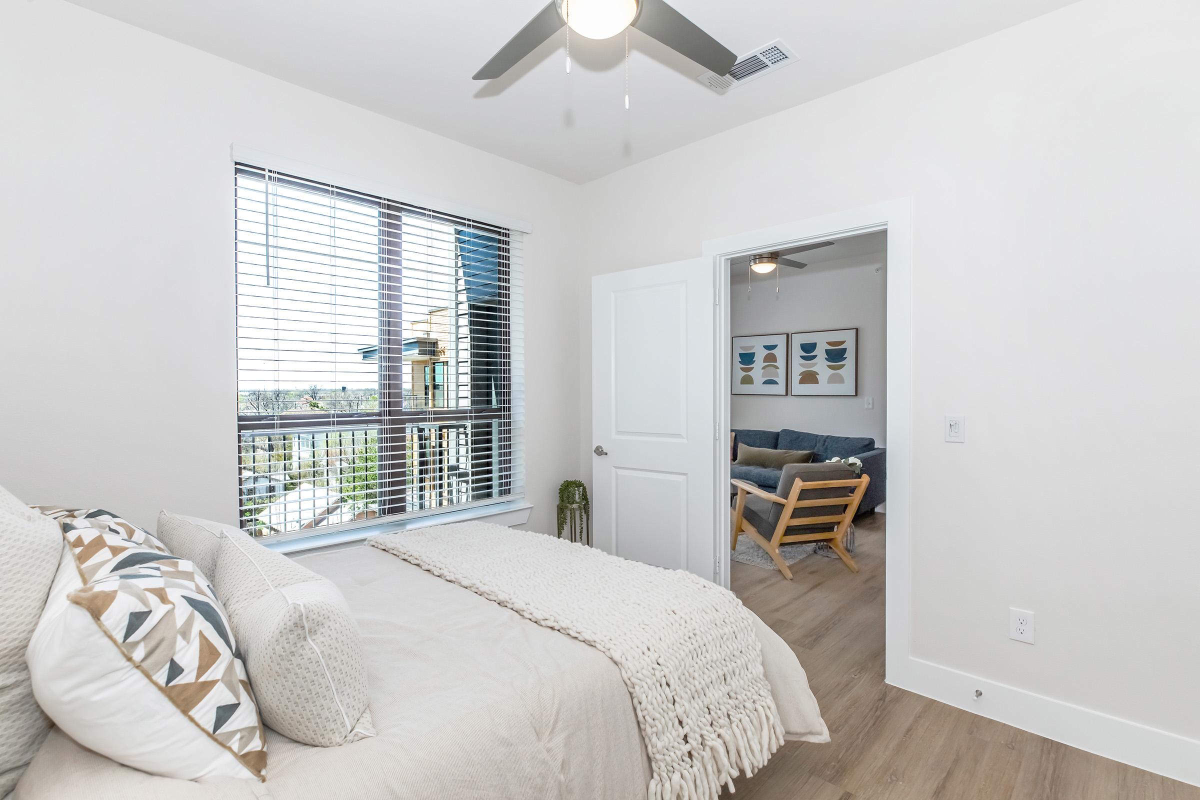 A modern bedroom with a neatly made bed featuring decorative pillows and a knitted throw. There is a large window with blinds allowing natural light. A door to another room is visible, and in the background, a living area is partially visible with a couch and artwork on the wall. The floor is light wood.