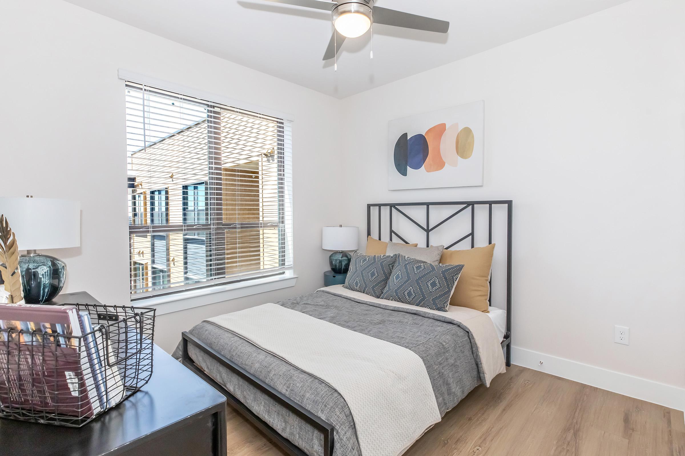 A cozy bedroom featuring a modern bed with gray and yellow bedding, two bedside lamps, and a decorative wall art piece. Natural light filters through a window with blinds, illuminating the wooden floor and adding a warm atmosphere to the space. A stylish basket sits on a dark bedside table.