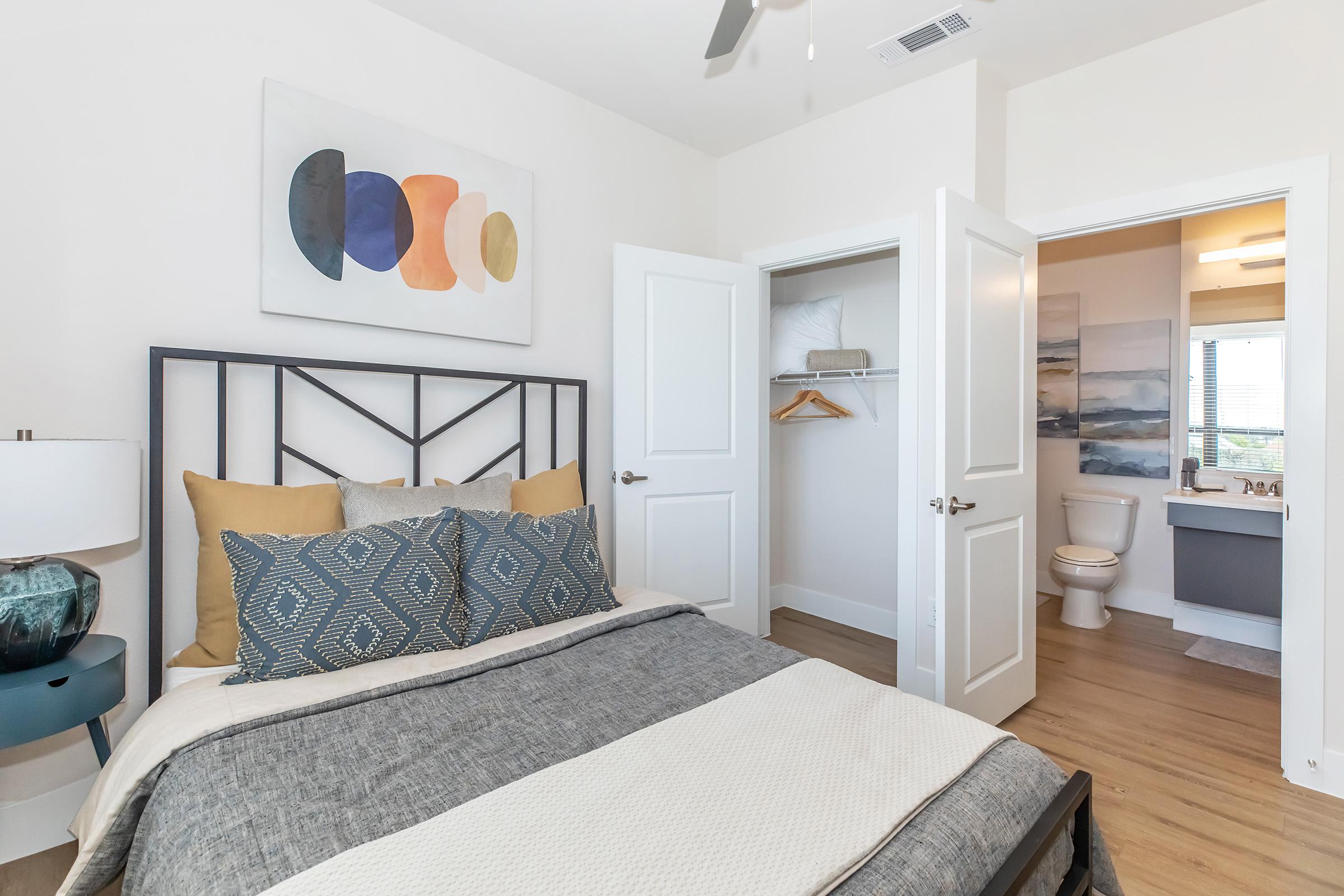 A modern bedroom featuring a bed with decorative pillows and a neutral-colored throw blanket. A wall art piece hangs above the bed, and there's a bedside table with a lamp. A doorway leads to a bathroom, visible in the background, which includes a toilet and sink area. Natural light comes from a window.