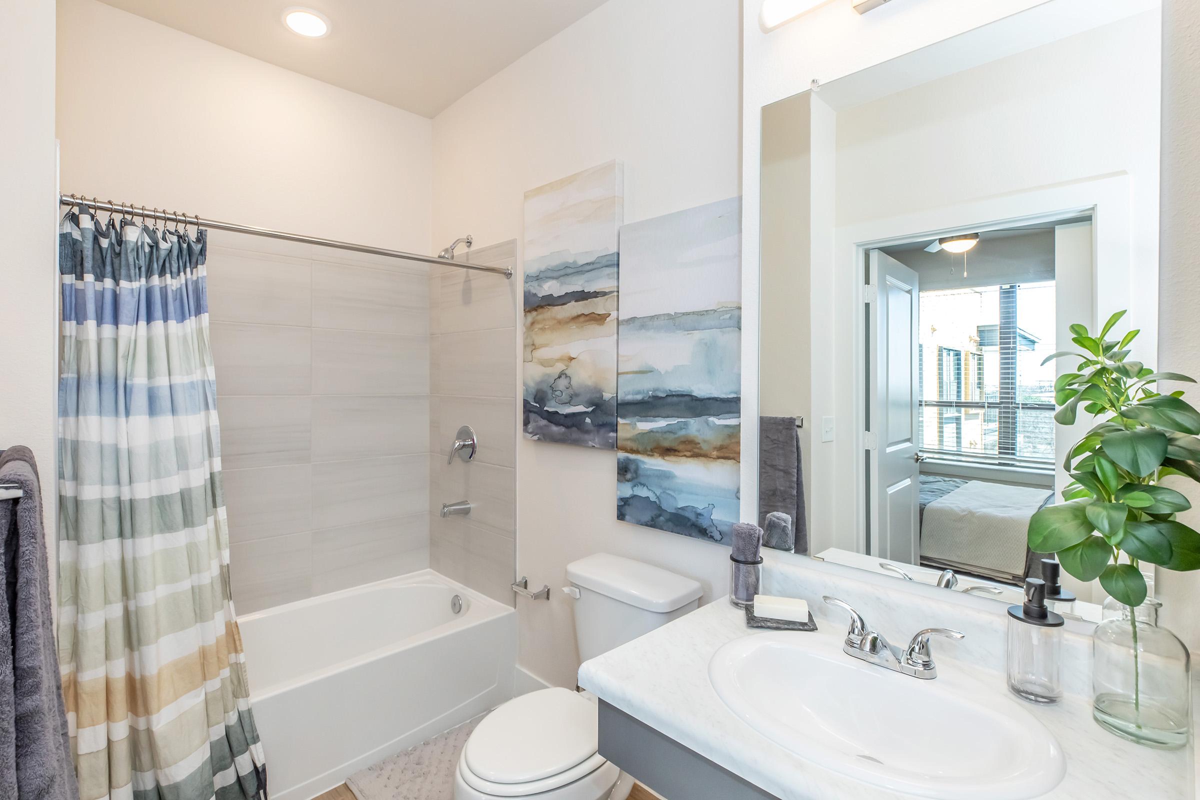 A modern bathroom featuring a bathtub with a shower curtain, a sleek sink with toiletries, and a large mirror. Soft, neutral colors dominate the decor, complemented by two abstract paintings on the wall. A leafy plant adds a touch of greenery, while a door leads to an adjoining room.