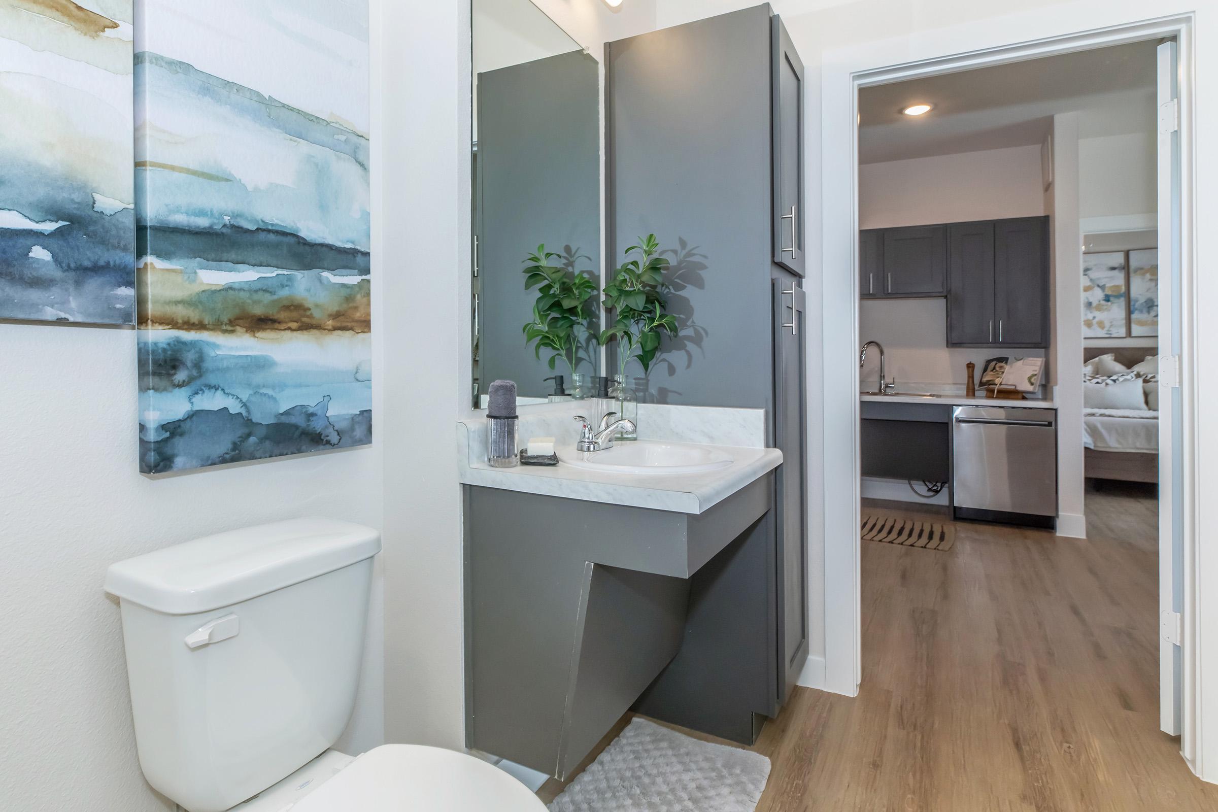 A modern bathroom featuring a white toilet, a sleek sink with a marble countertop, and stylish cabinetry. There's a small potted plant on the countertop and a watercolor painting on the wall. The image also shows part of a kitchen in the background, with dark cabinetry and a glimpse of a living space.