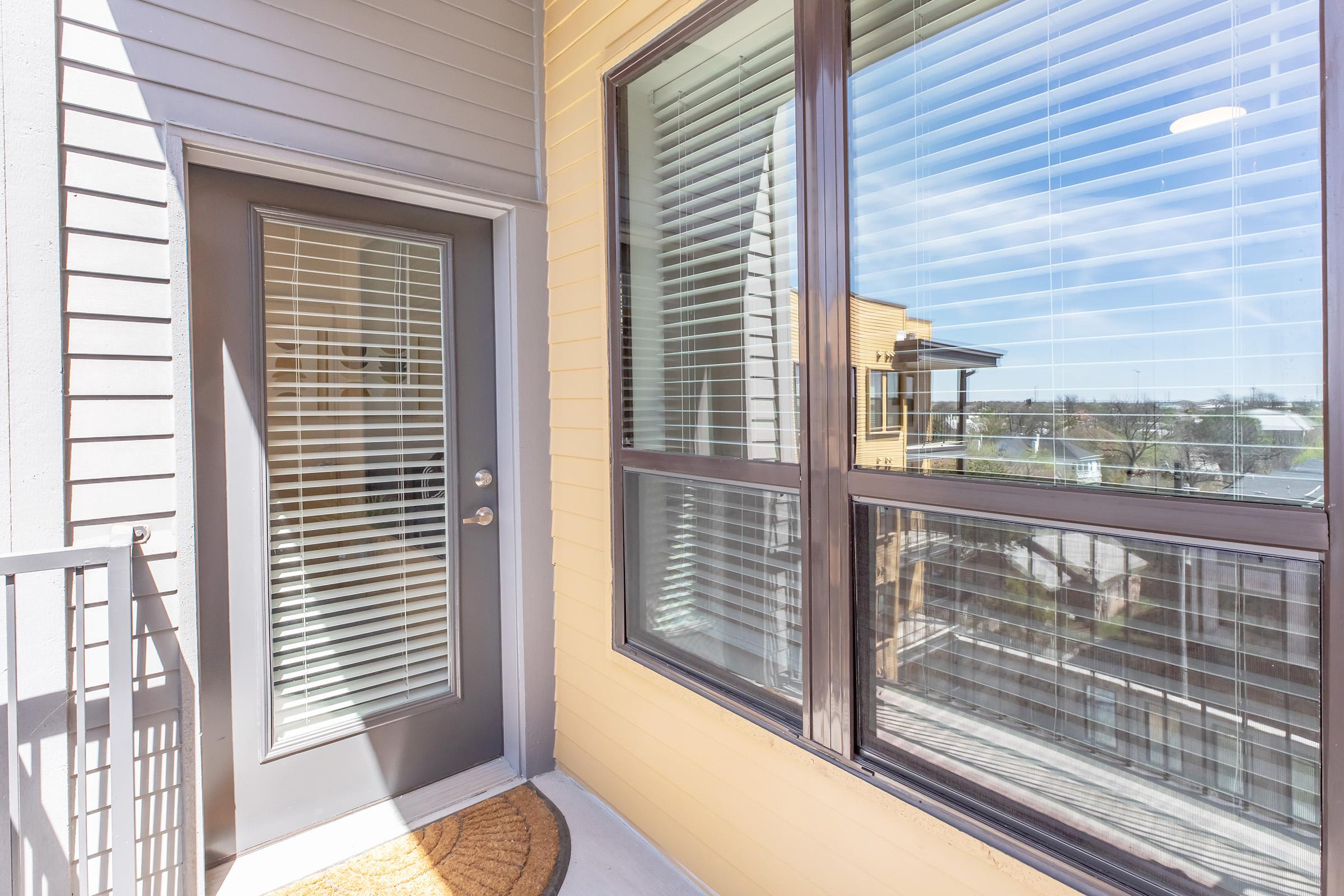 A well-lit balcony featuring a door with horizontal blinds and large windows. The exterior walls are a mix of gray and yellow siding. A doormat is positioned at the entrance, and the view outside includes rooftops against a clear blue sky.