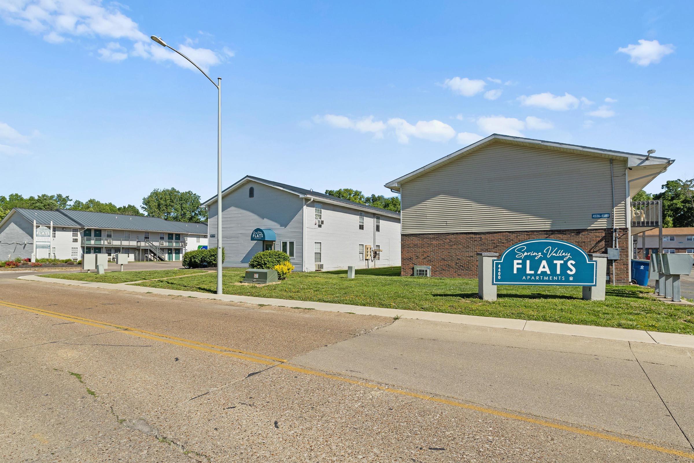A view of Spring Village Flats Apartments, featuring multiple residential buildings with a sign indicating the complex. The area has landscaped greenery and a paved road in the foreground, with blue skies and clouds in the background. The setting appears to be well-maintained and welcoming.