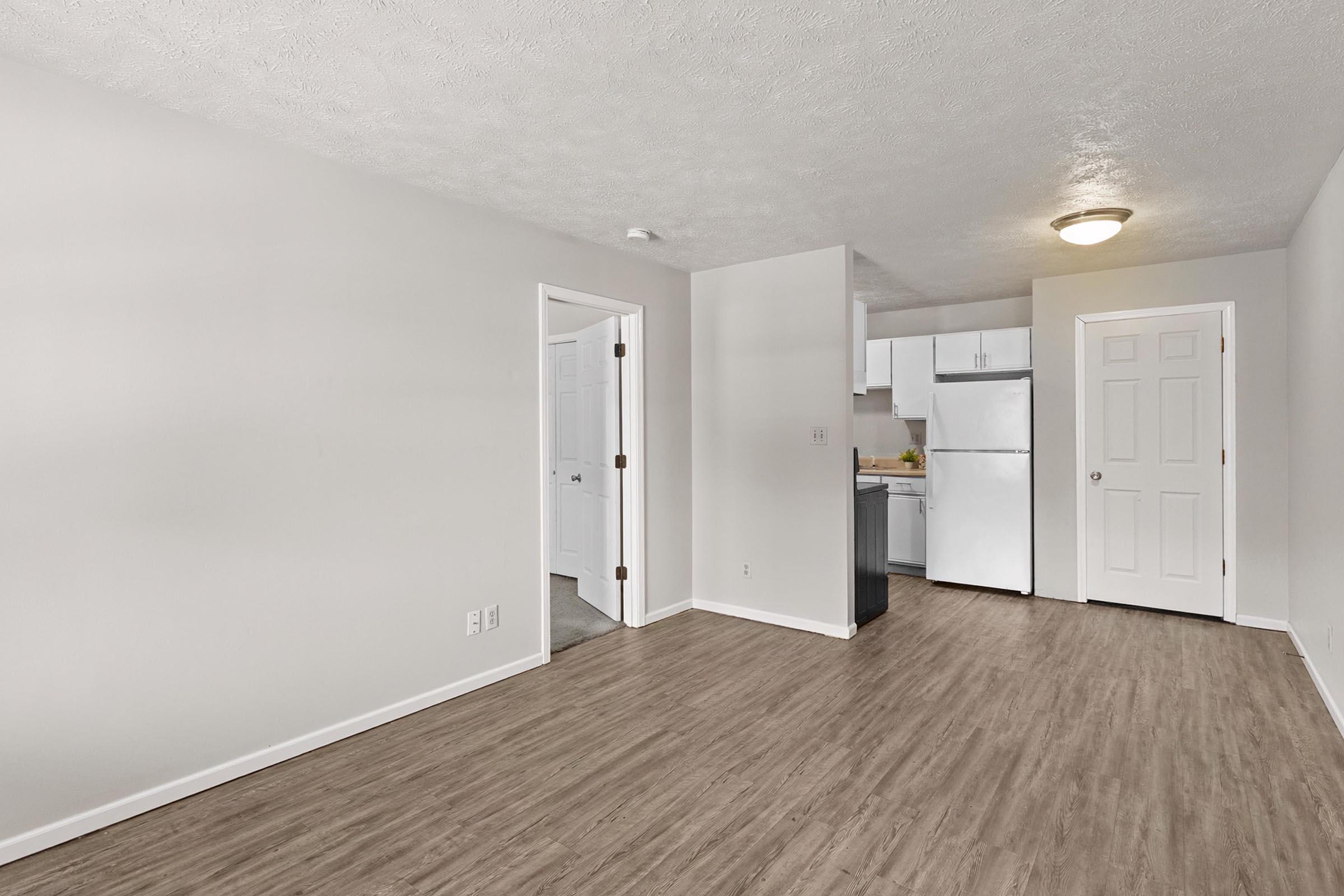 A modern, light-filled apartment interior featuring an open living area with laminate flooring. To the left, a white door leads to another room, while the kitchen is visible in the background with white appliances and cabinetry. The walls are neutral-colored, enhancing the spacious feel of the environment.