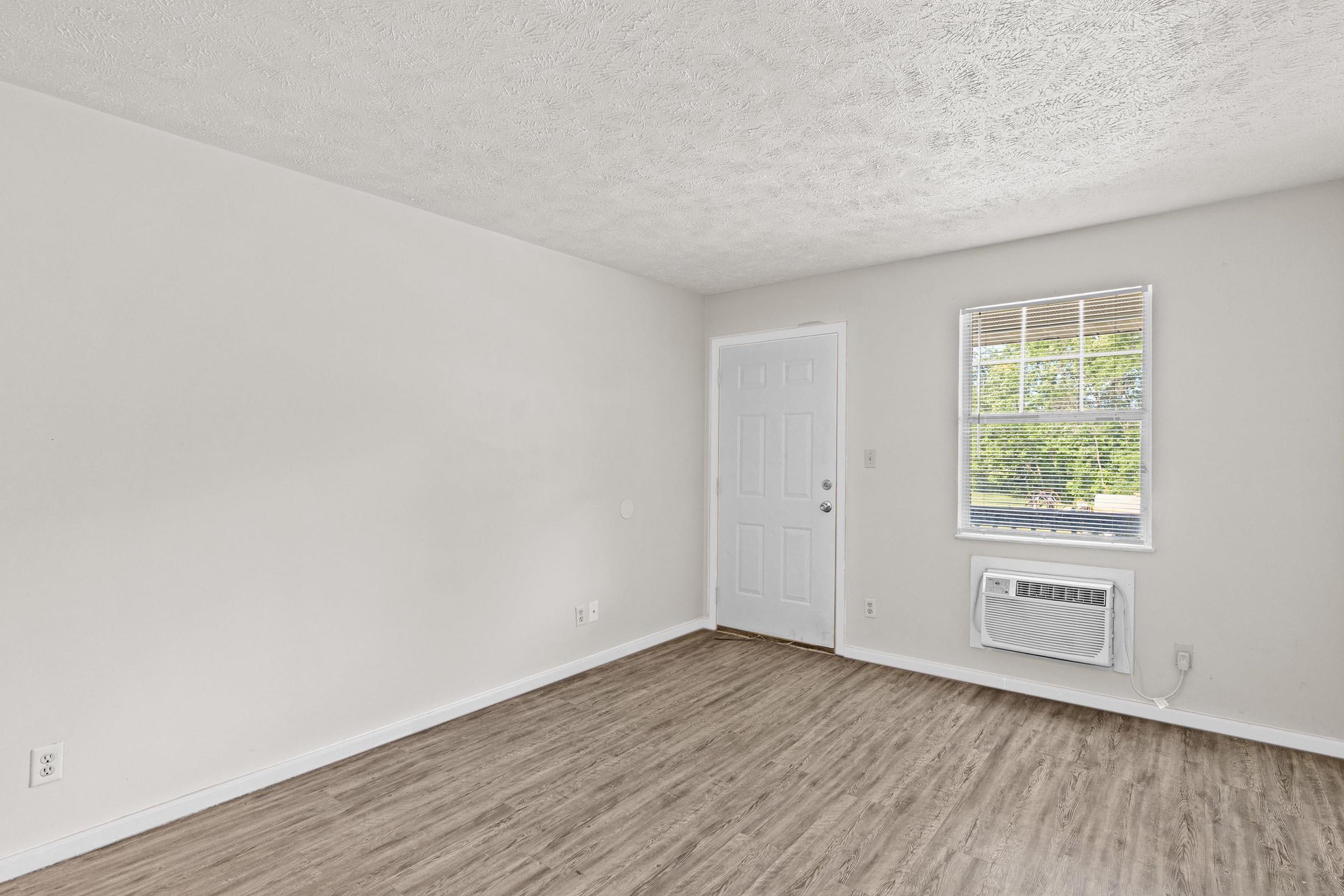 An empty room featuring light-colored walls and a large window with blinds. There is an air conditioning unit beneath the window and a plain door on one side. The floor has wood-like vinyl, creating a clean and spacious appearance with natural light streaming in.