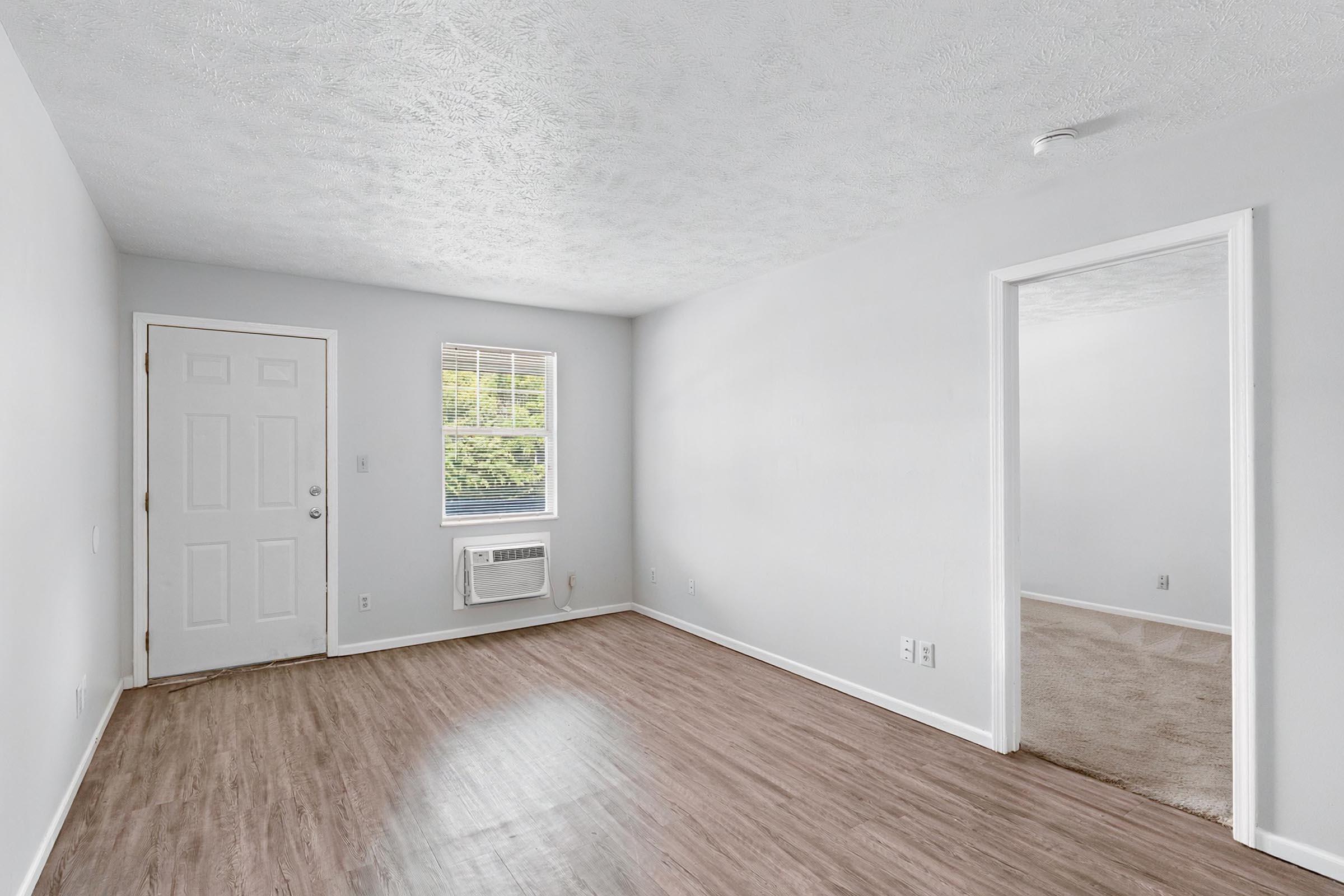 A spacious, empty room with light gray walls and wooden laminate flooring. There is a white door leading to the outside, a window with blinds providing natural light, and an air conditioning unit mounted on the wall. A doorway on the right leads to another room.
