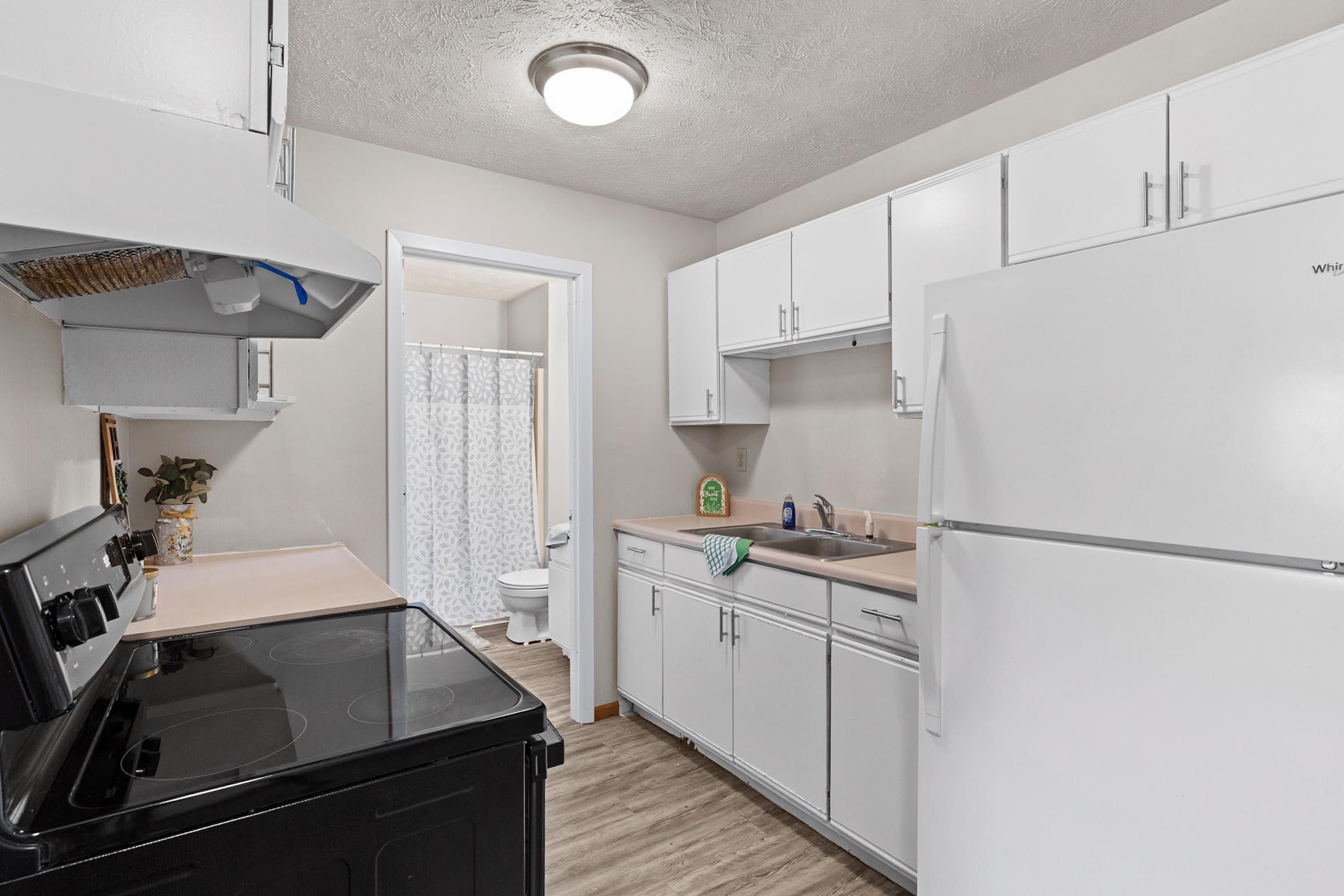 A clean, modern kitchen featuring white cabinets, a black stove, and a refrigerator. The countertop is light pink, and there's a small sink. A doorway leads to a bathroom, visible in the background, with a shower curtain and toilet. Natural light brightens the space, enhancing the inviting atmosphere.