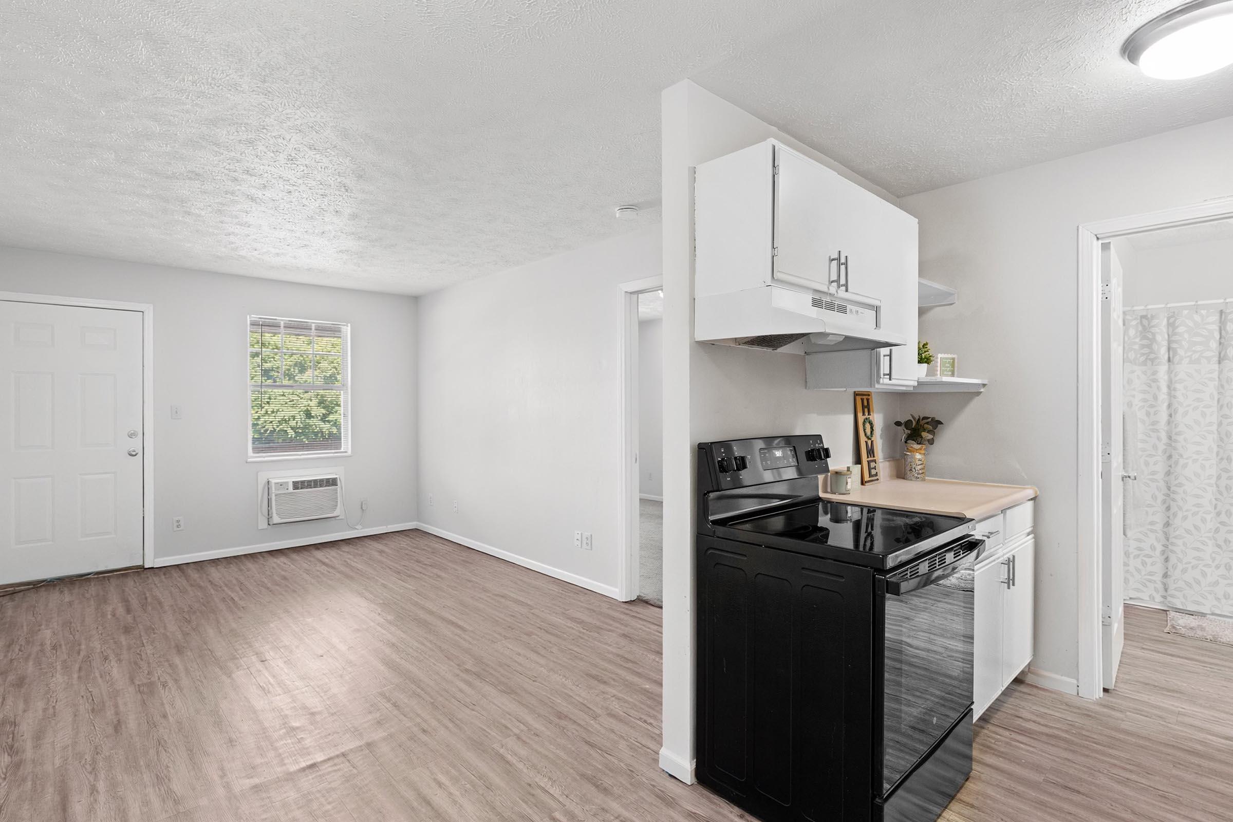 Interior view of a modern, bright apartment featuring an open living space. On the left, there's a front door and a window allowing natural light. To the right, a kitchen area with black appliances, including a stove and dishwasher, and a countertop. Light-colored walls and wood-style flooring enhance the spacious feel.