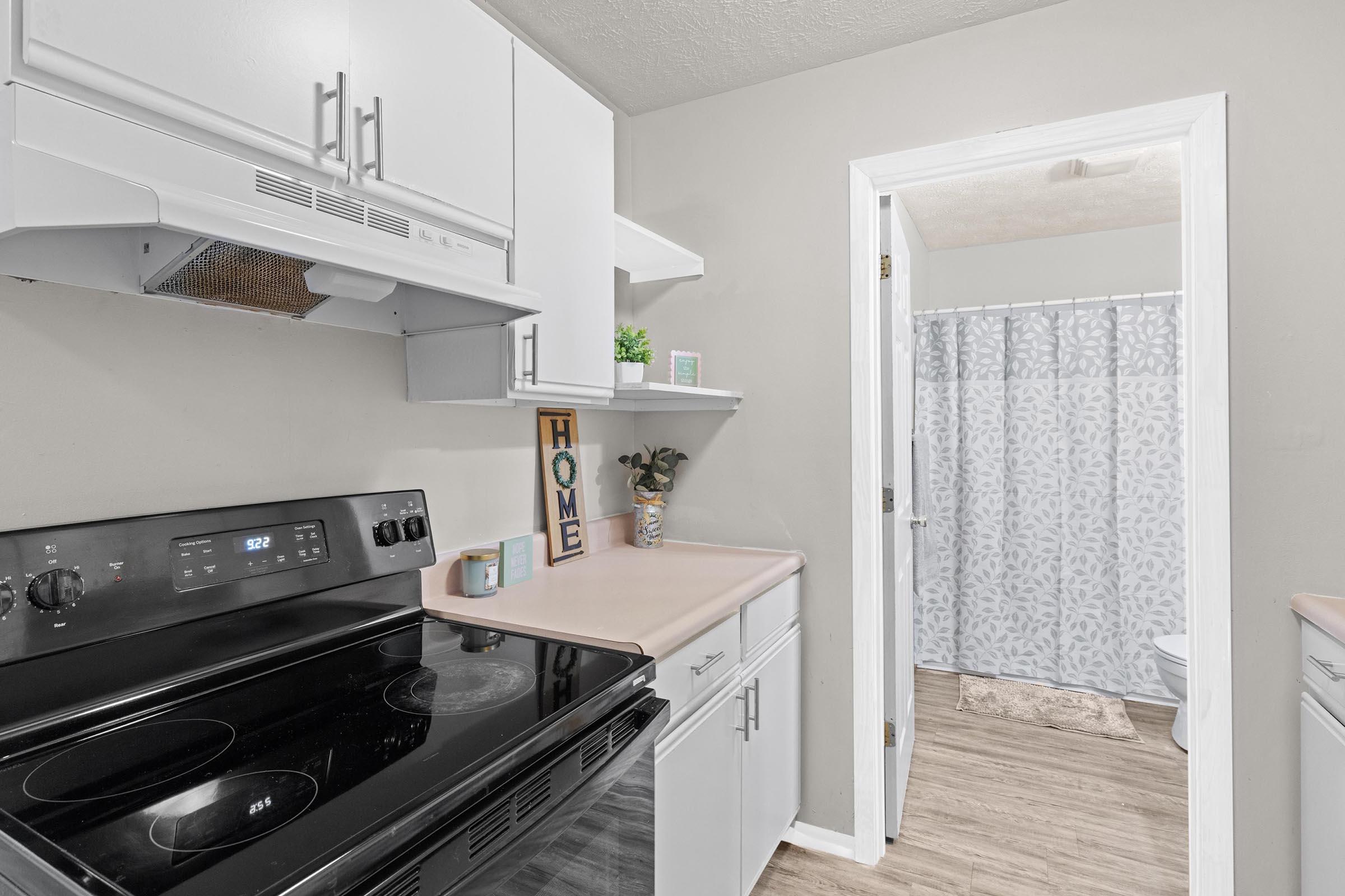 Modern kitchen with white cabinets and a black stove, featuring a light pink countertop. A small plant and decorative sign are on the counter. In the background, a bathroom doorway with a gray patterned shower curtain is visible. The walls are painted a neutral shade, enhancing the overall brightness of the space.