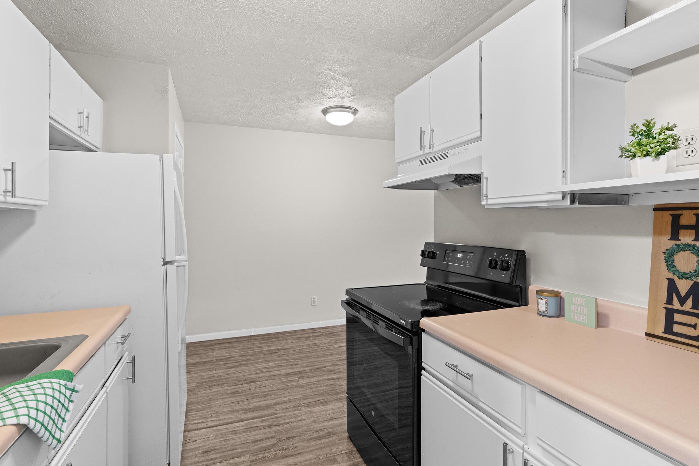 A bright, modern kitchen featuring white cabinets, a black stove, and a refrigerator. The countertops are light-colored, with a sink on the left and an oven on the right. There's a green kitchen towel and a decorative sign that says "Home" on the counter. The space has a clean, inviting atmosphere.