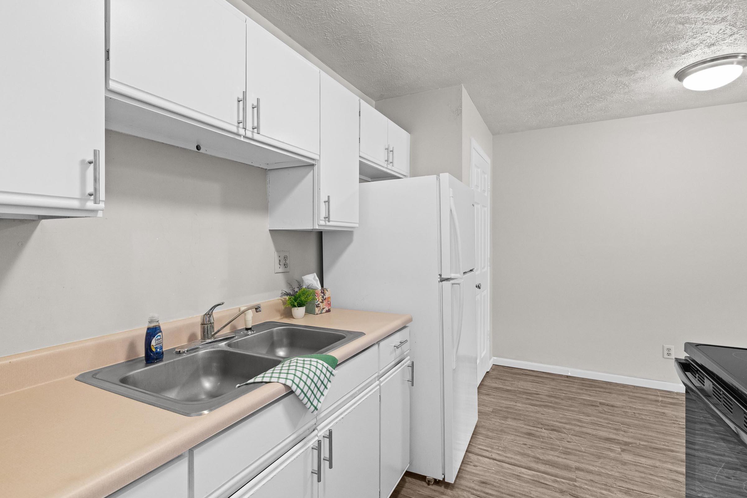 A clean and organized kitchen with white cabinets, a double sink, and a green checkered dish towel. The countertop is beige, and there is a potted plant near the sink. A white refrigerator stands against the wall, and the flooring is a light wood laminate. The walls are painted a neutral color.