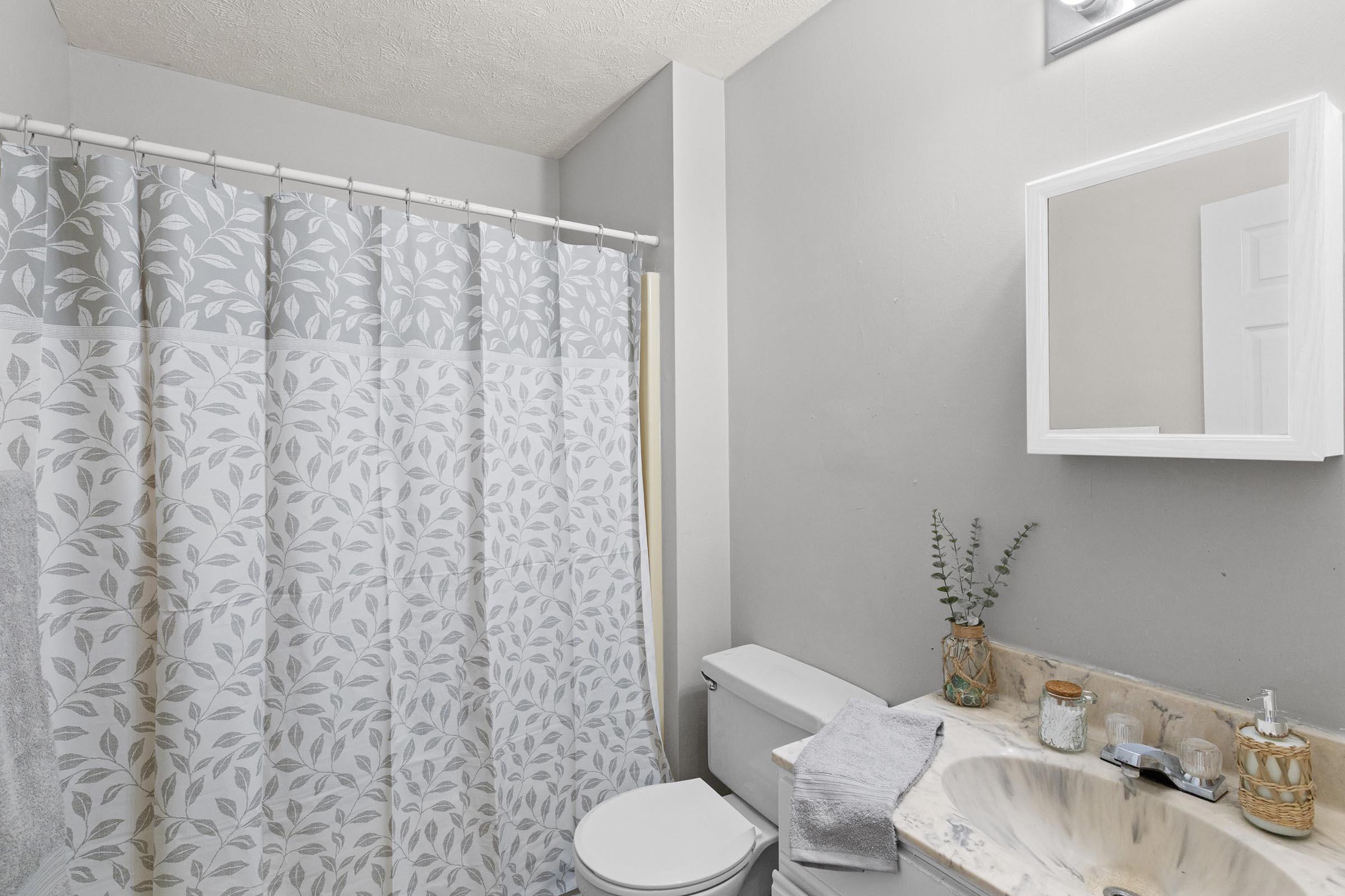 A clean and modern bathroom featuring a shower with a leaf-patterned curtain, a white toilet, and a marble sink countertop. A small vase with greenery is placed next to the sink, and a mirror hangs above it. The walls are painted light gray, creating a serene atmosphere.