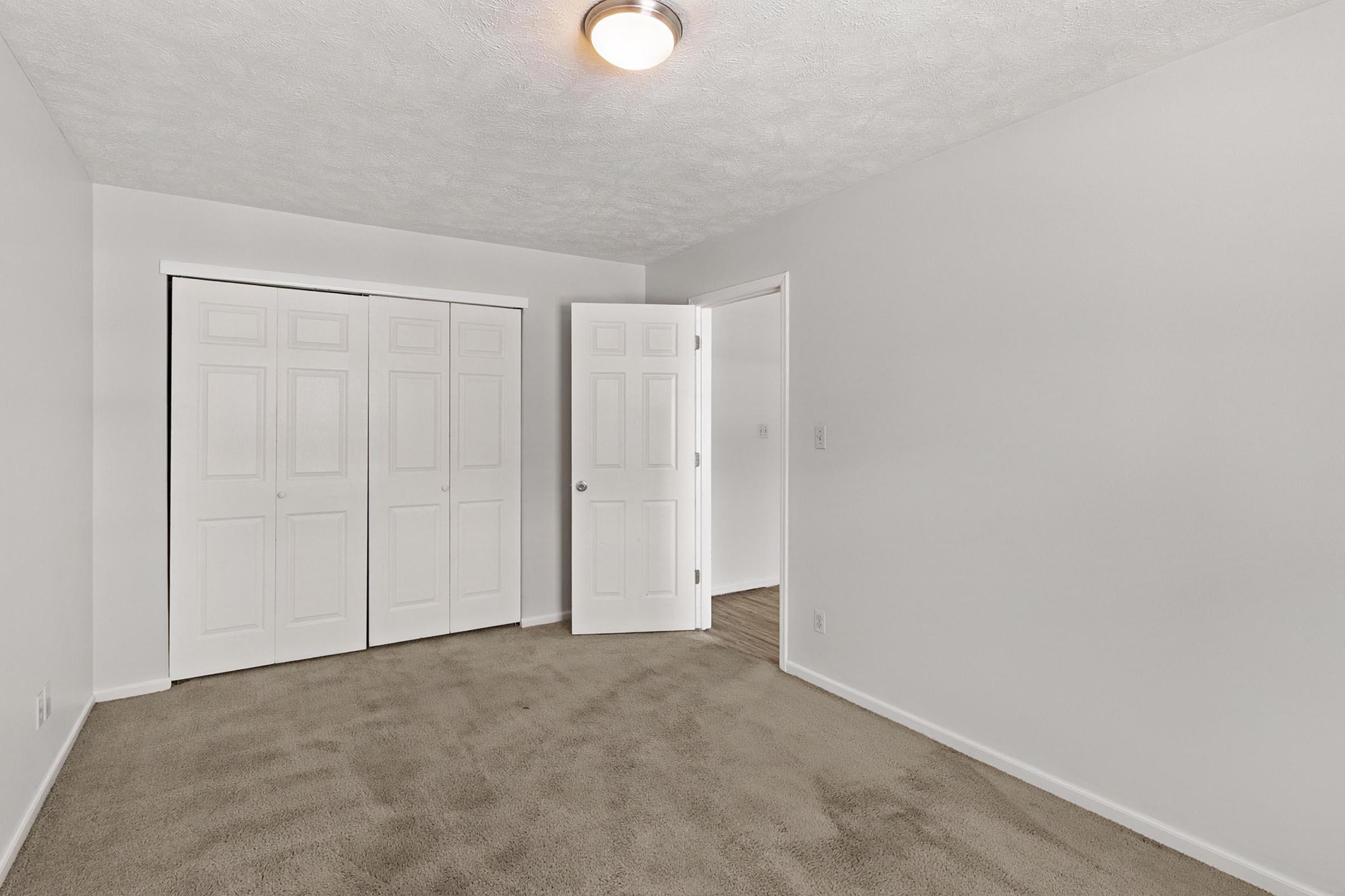 Empty room with light-colored walls and carpet. Two open white doors reveal sliding closet doors. There is a ceiling light fixture, and the floor is clean and undisturbed, giving a spacious feel.
