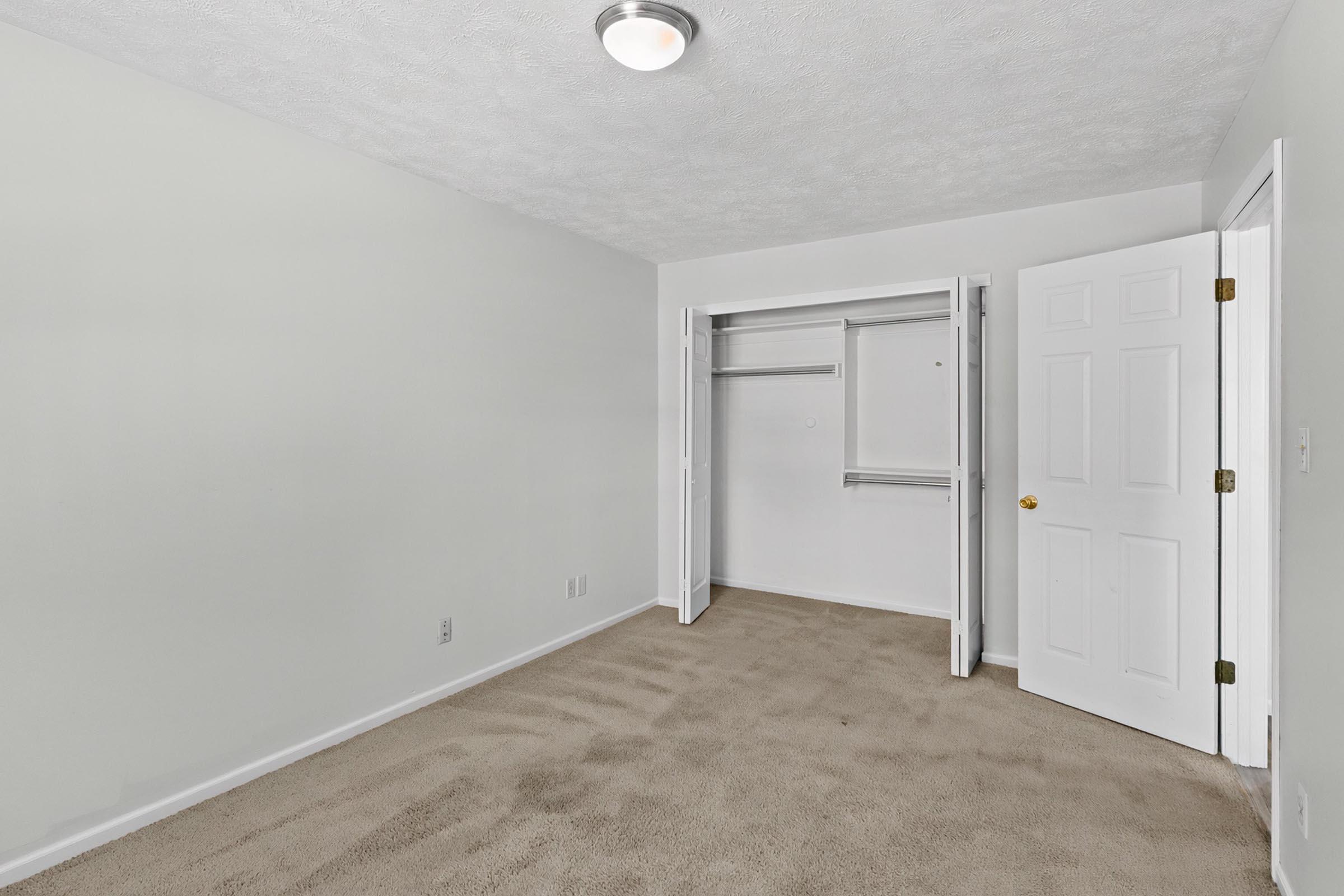 Empty bedroom with light-colored walls and carpeted floor. A closet with sliding doors is on the left, and a door is partially open on the right, leading to another space. The ceiling has a light fixture, and the room appears clean and spacious, ready for furnishing.