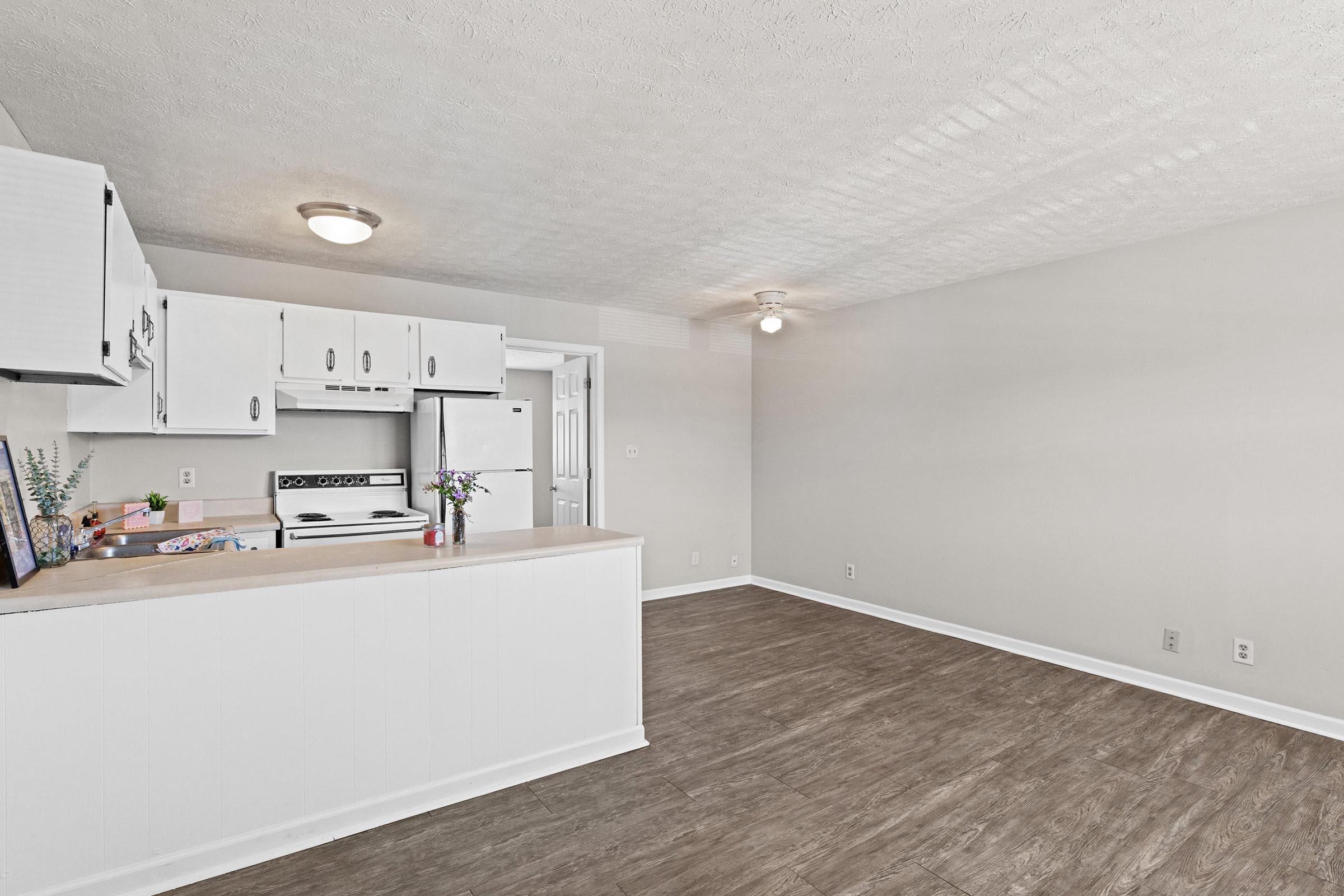 Interior of an apartment featuring a kitchen with white cabinets, a refrigerator, and an open floor plan. The living area has light-colored walls and wooden flooring, with a ceiling light fixture. A small table with decorative items is visible in the kitchen area. Overall, the space appears bright and inviting.